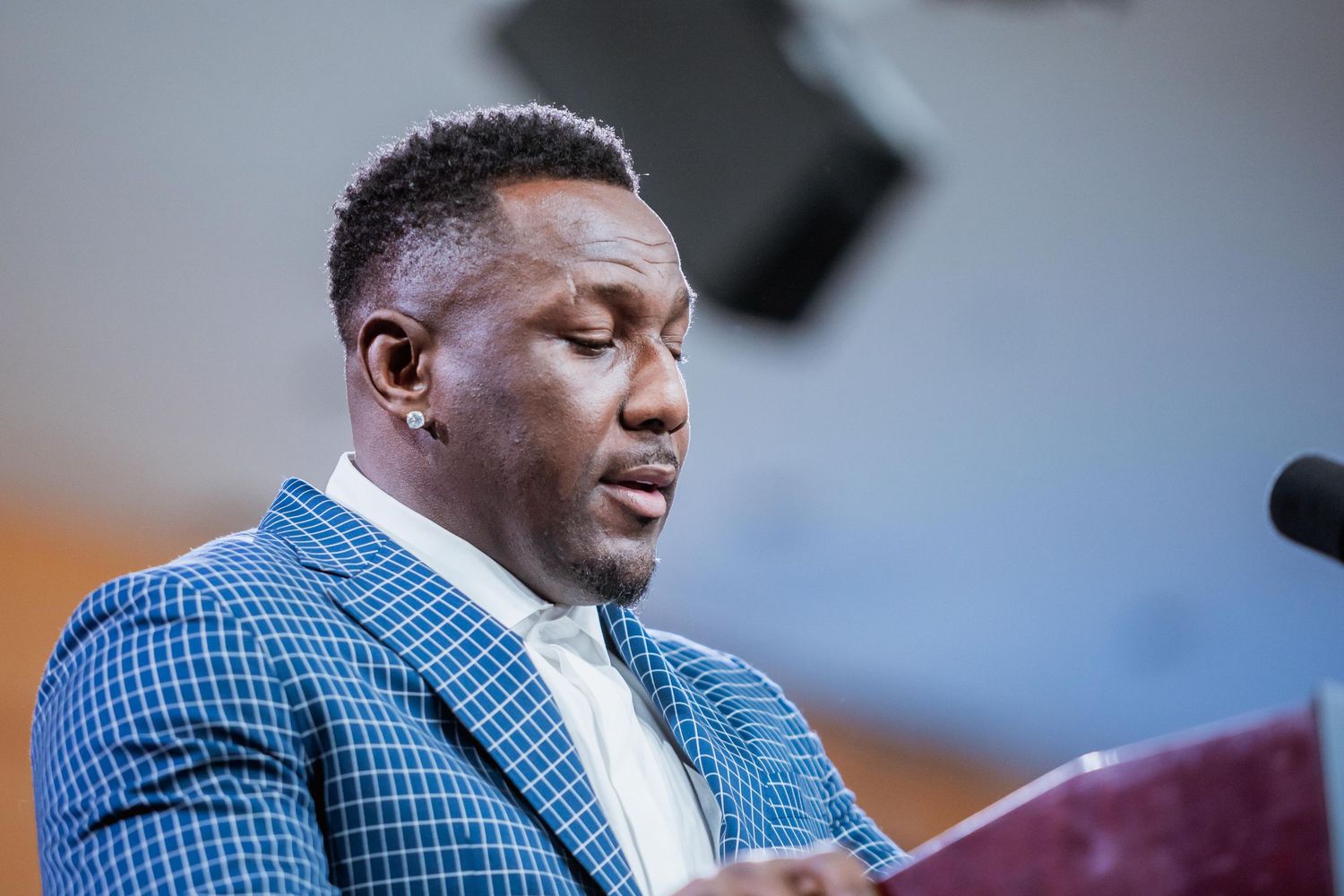 A man in a blue checkered suit speaks at a podium during a formal event.