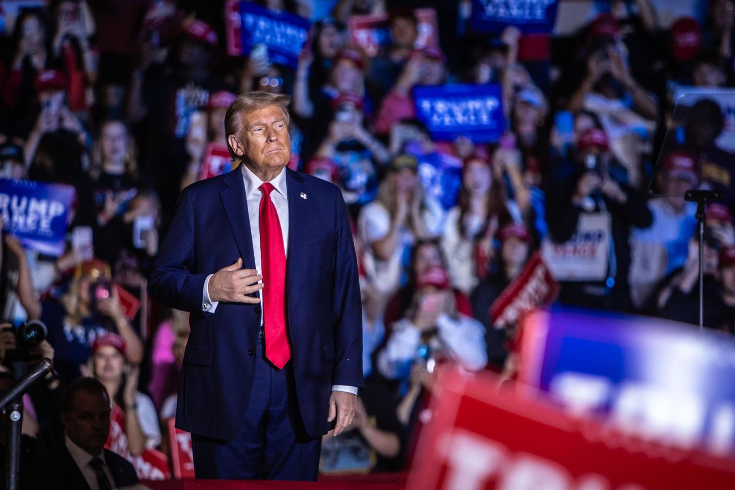 Donald Trump stands confidently on stage at a packed campaign rally with supporters holding Trump Vance signs.