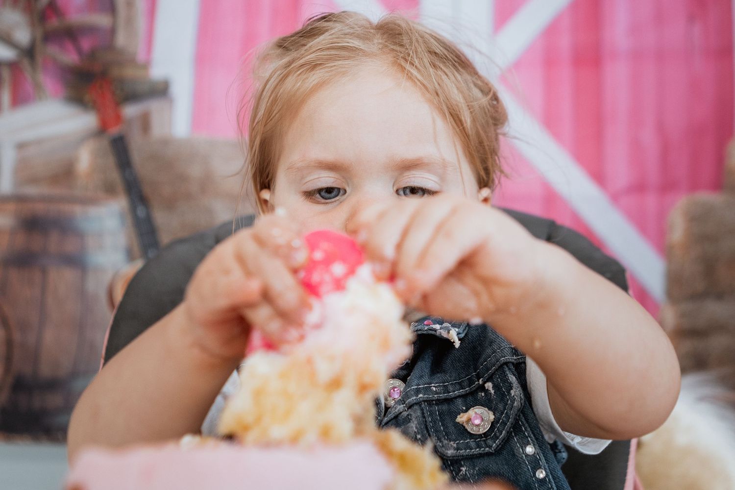 Young child in denim vest grabbing a pink decorated smash cake at a birthday party.