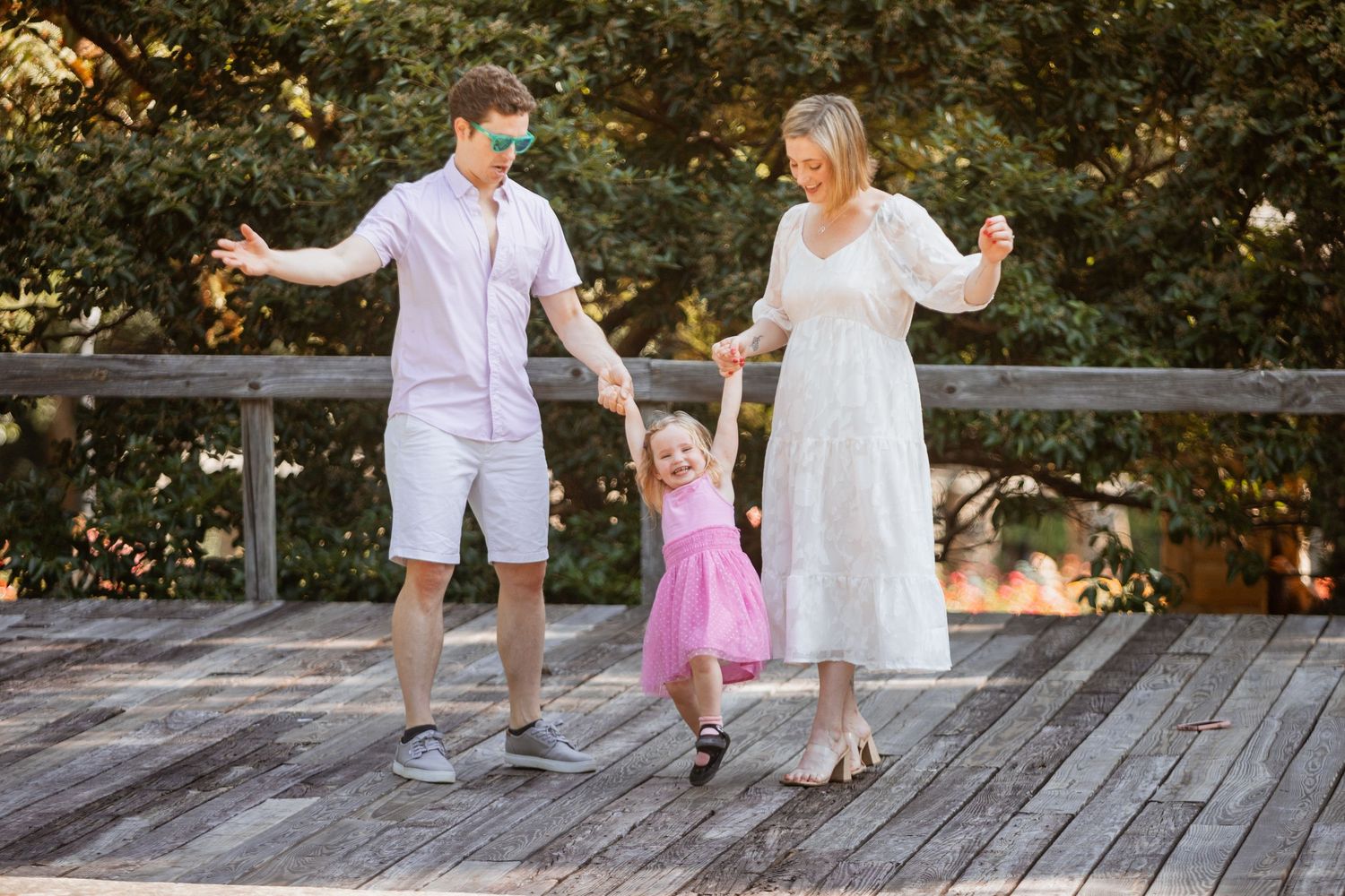 Joyful family swinging young daughter by hands on sunny wooden boardwalk in park.
