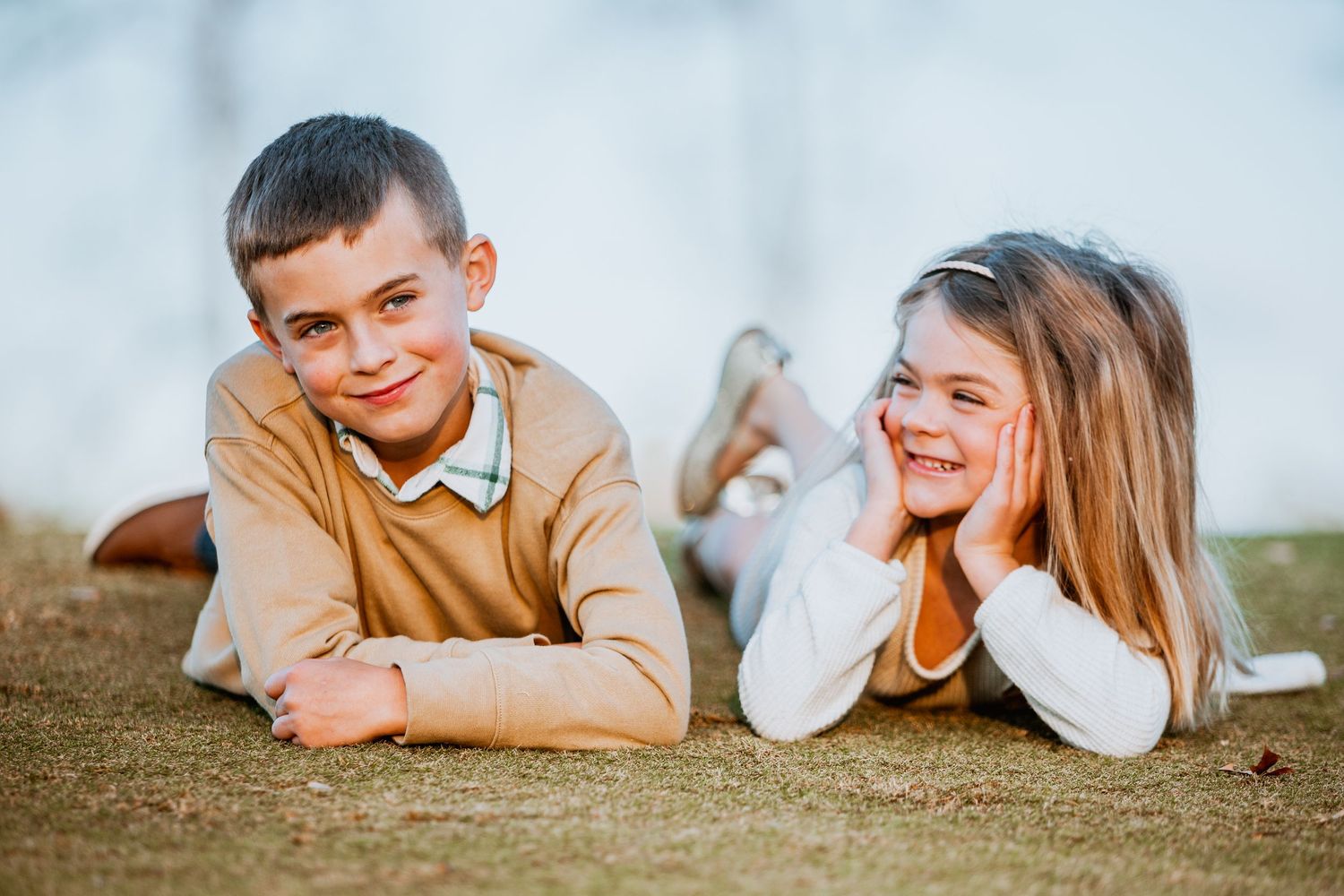 Boy and girl lying on grass laughing together during an outdoor family photo session in warm light.