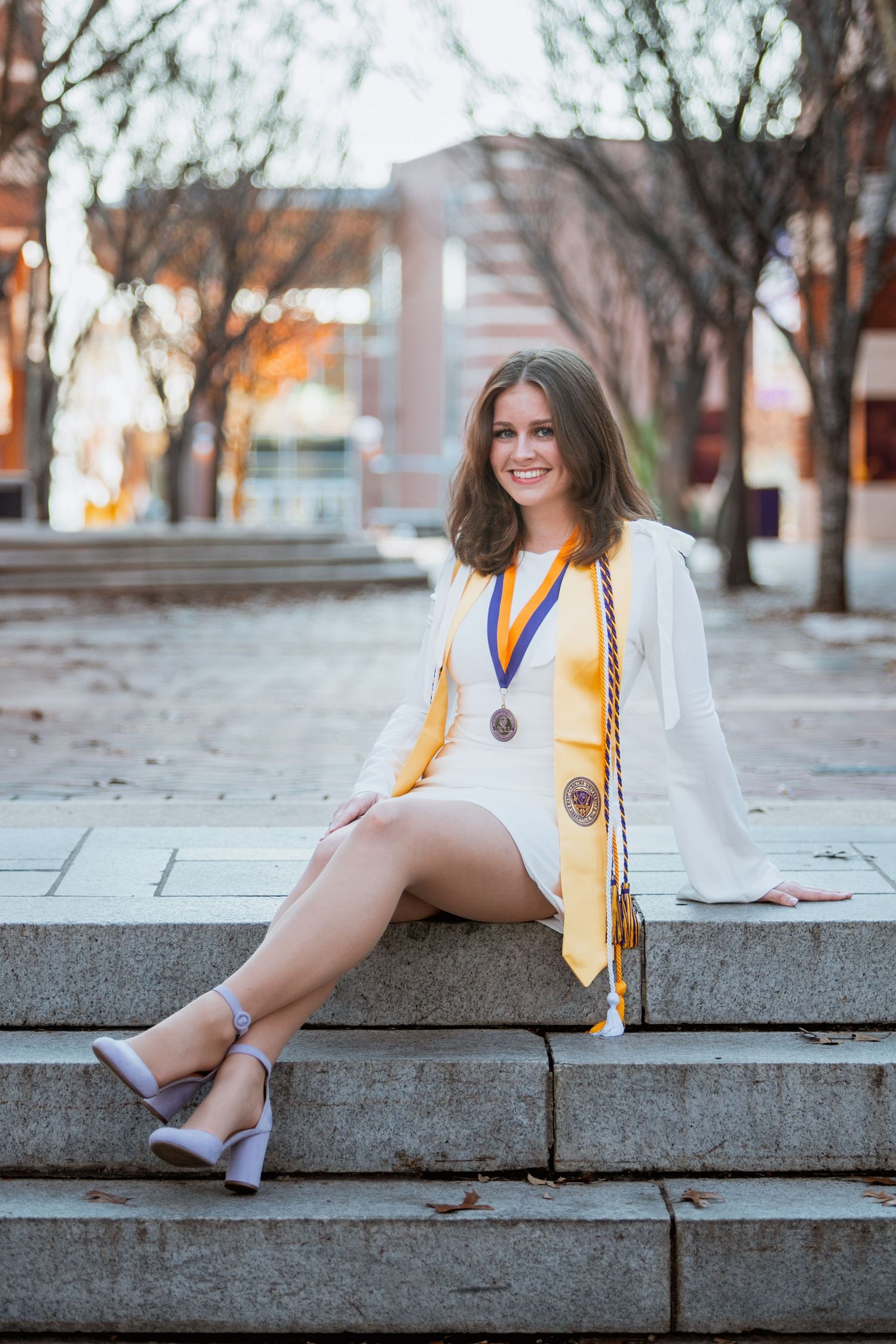 Young woman in white graduation dress with ECU gold stole and medal sits on stone steps smiling.