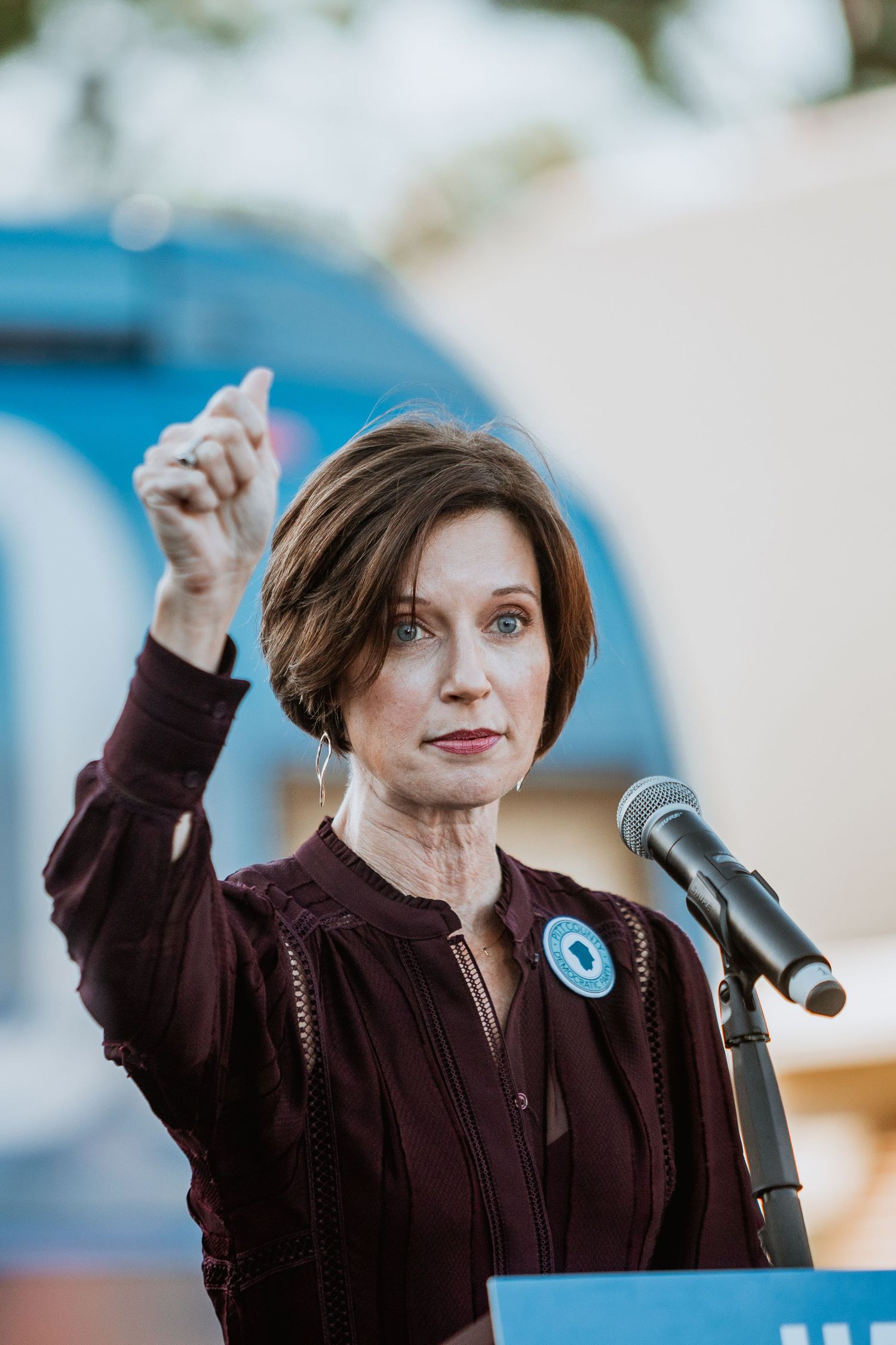 Woman with short brown hair speaking at a podium with a microphone, raising her finger outdoors.