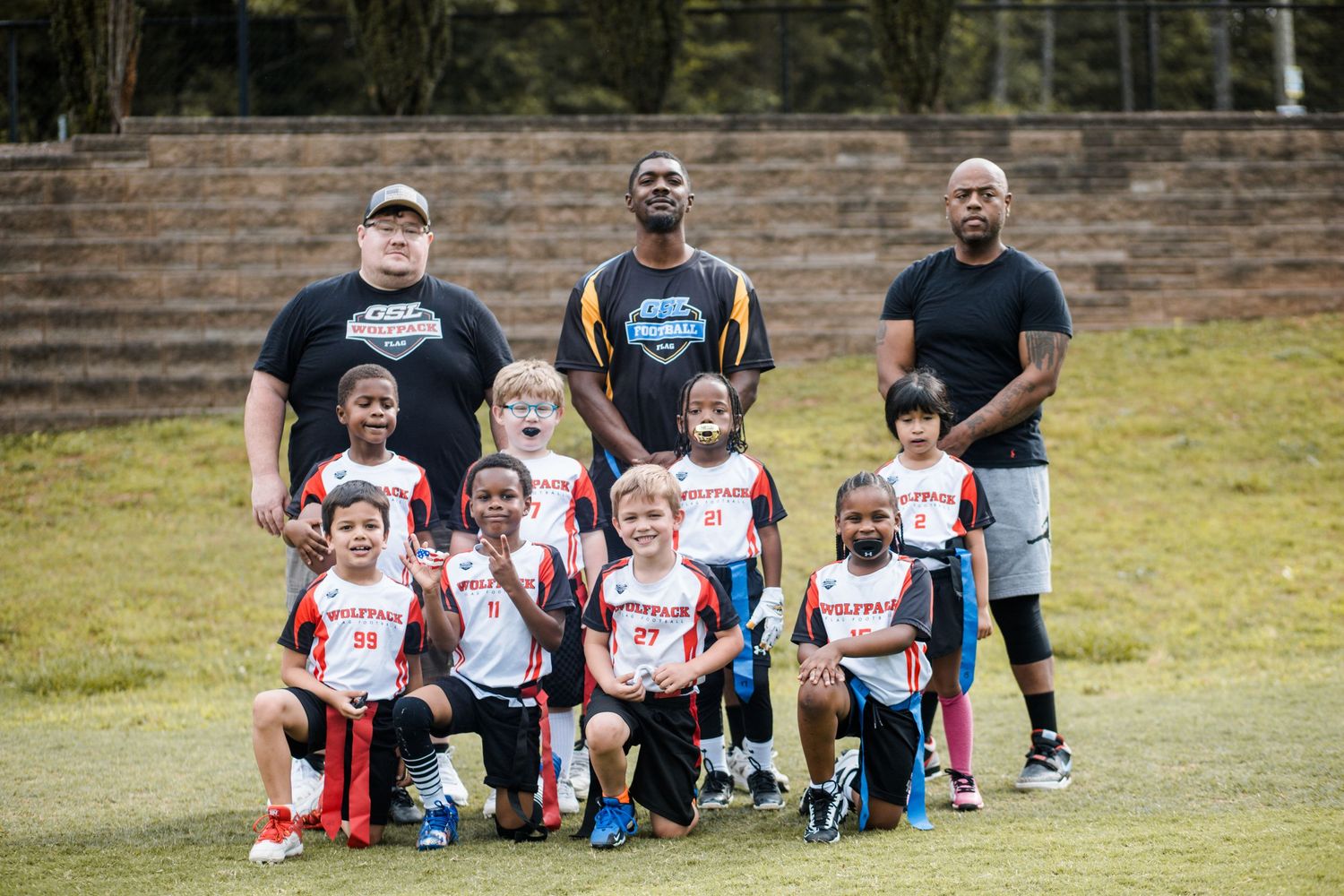 Youth football team in red and white jerseys poses with three adult coaches outdoors.