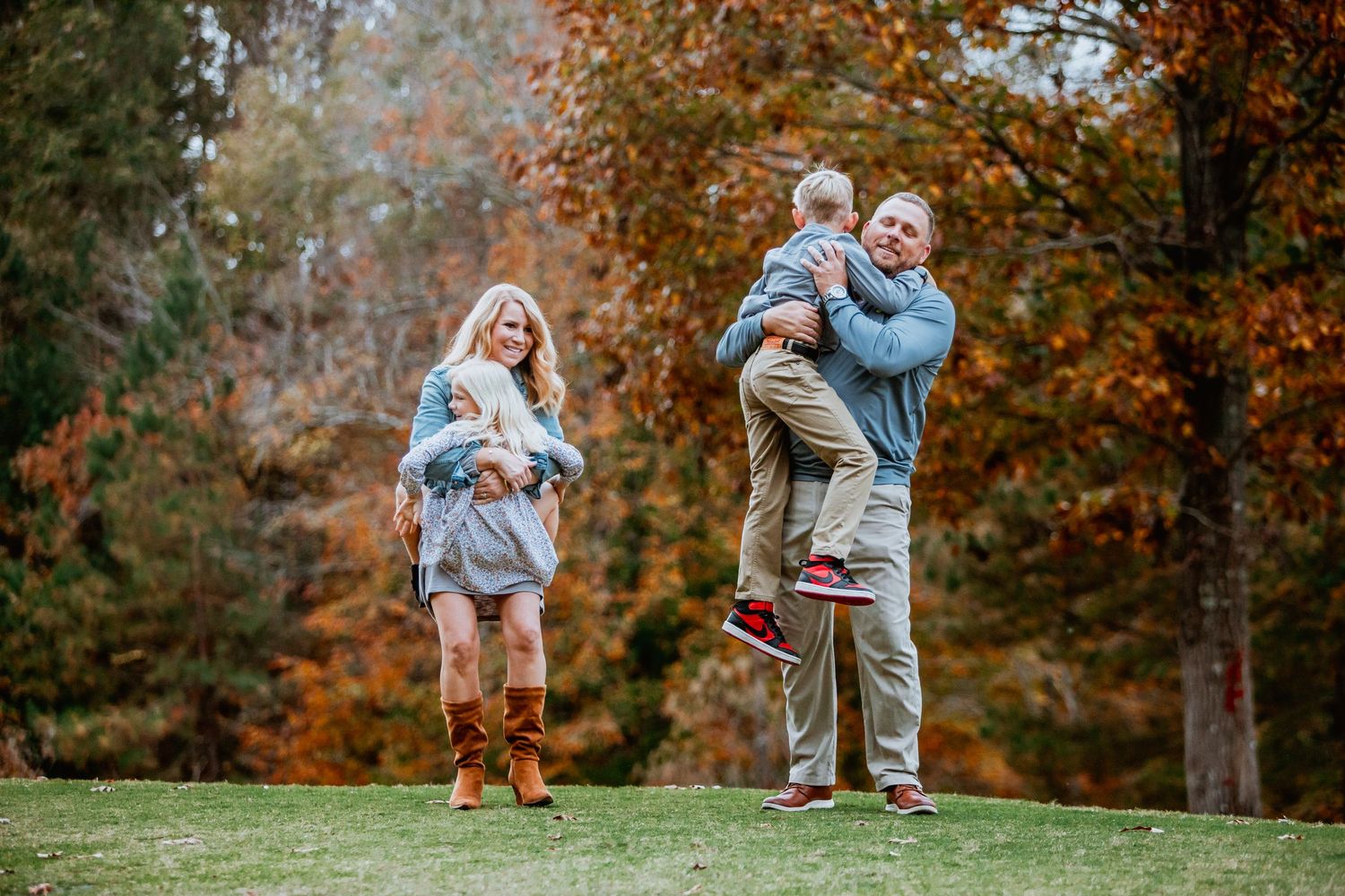 Family of four playing outdoors with parents lifting children amid vibrant fall foliage.