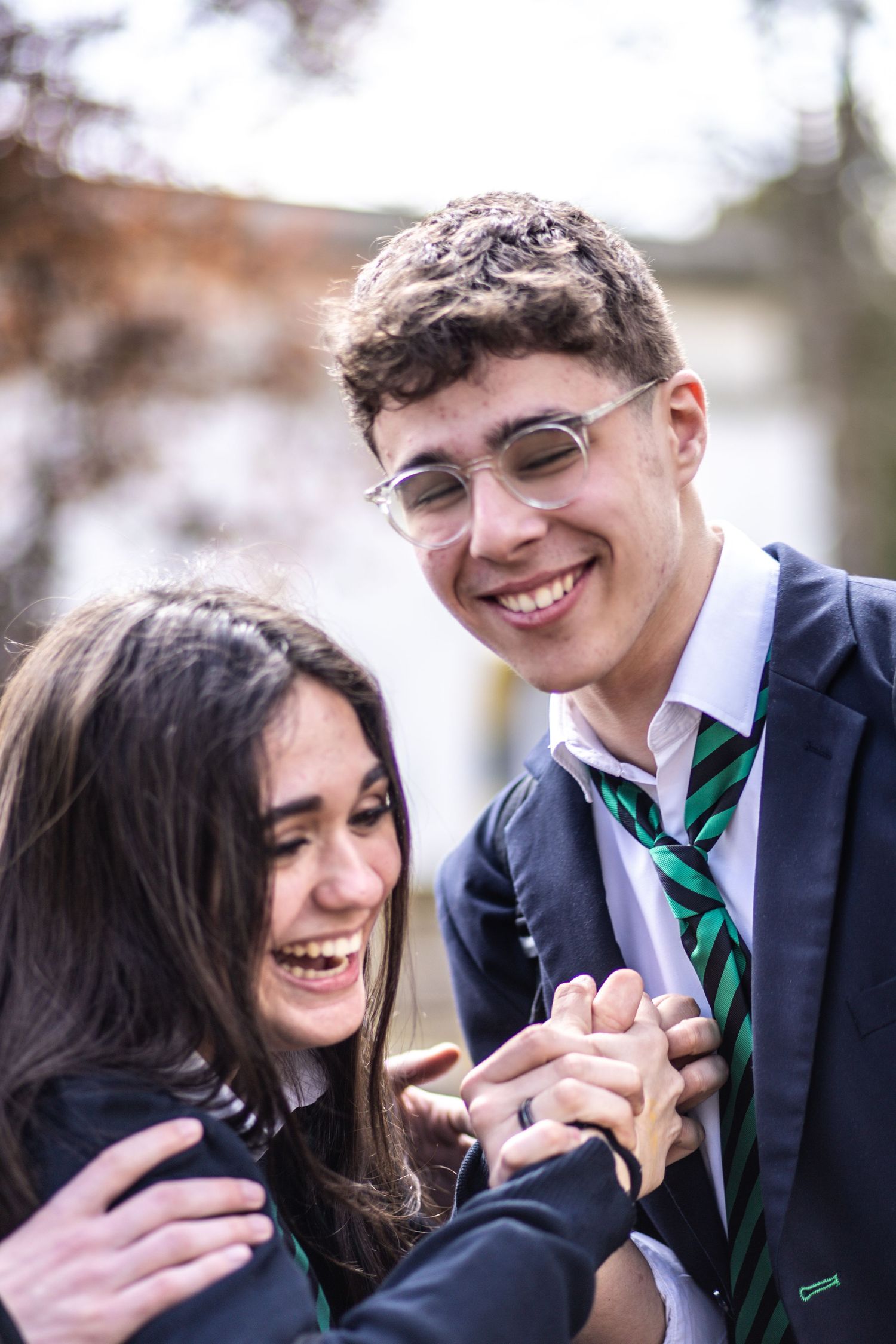 Two smiling teenagers in school uniforms with green striped ties laugh and hold hands outdoors.