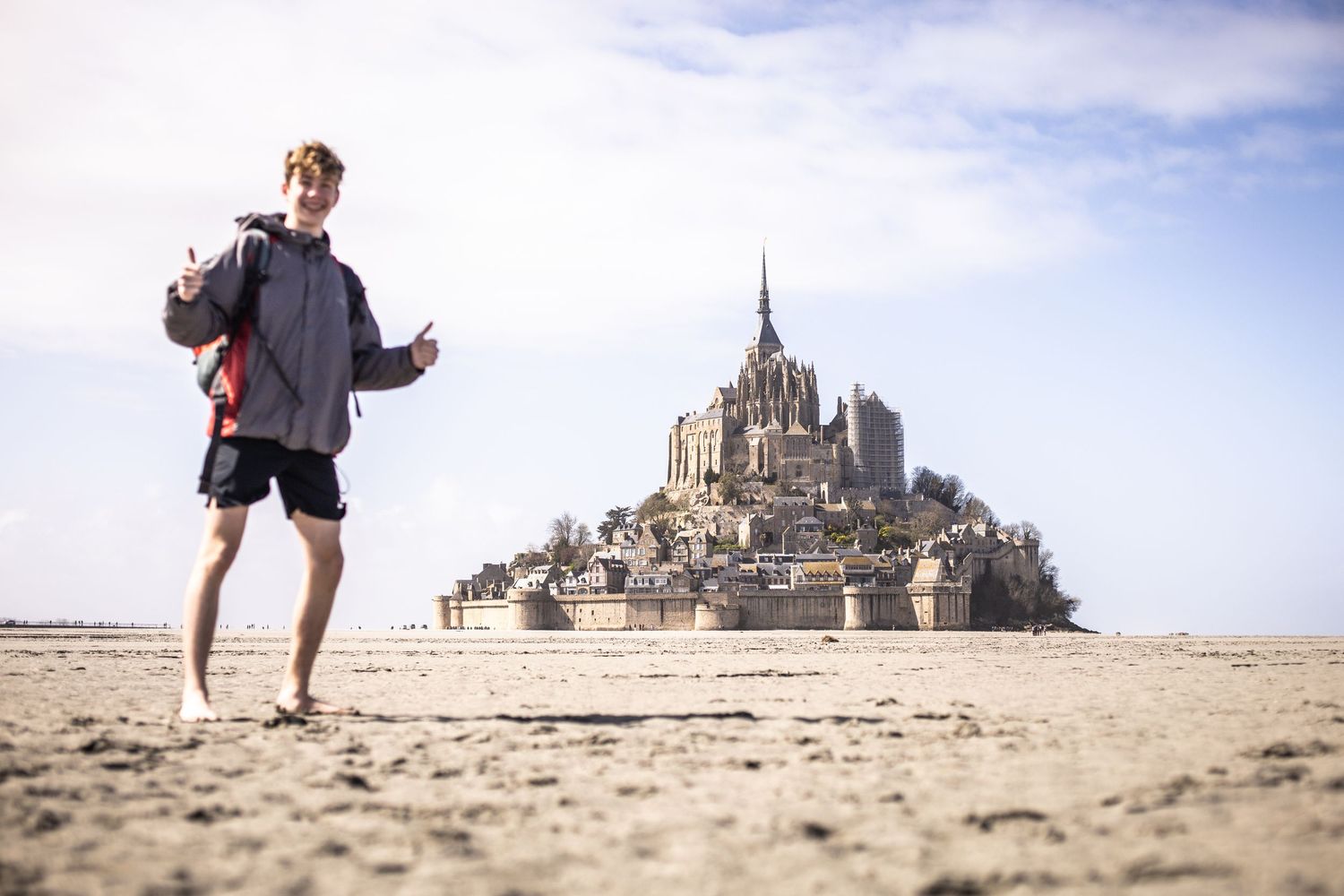 Young man walking on sandy tidal flats with iconic Mont Saint-Michel abbey in the background.