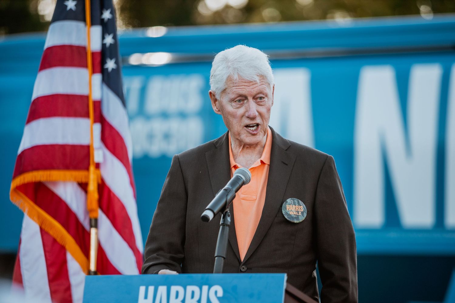 Man in brown jacket speaks at podium with Harris sign and American flag at outdoor political rally.