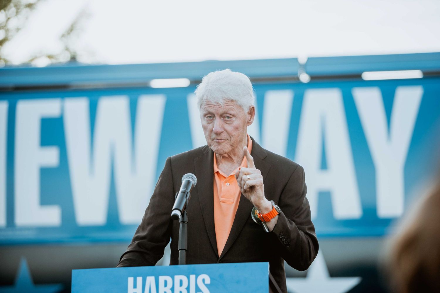 Elderly man in orange shirt speaks at Harris campaign podium with 'New Way' banner behind him.