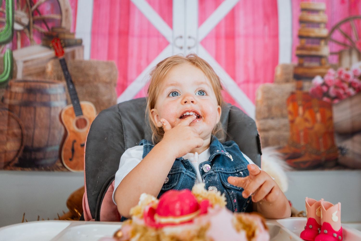 Adorable blonde toddler girl eating birthday cake at farm-themed first birthday party celebration.