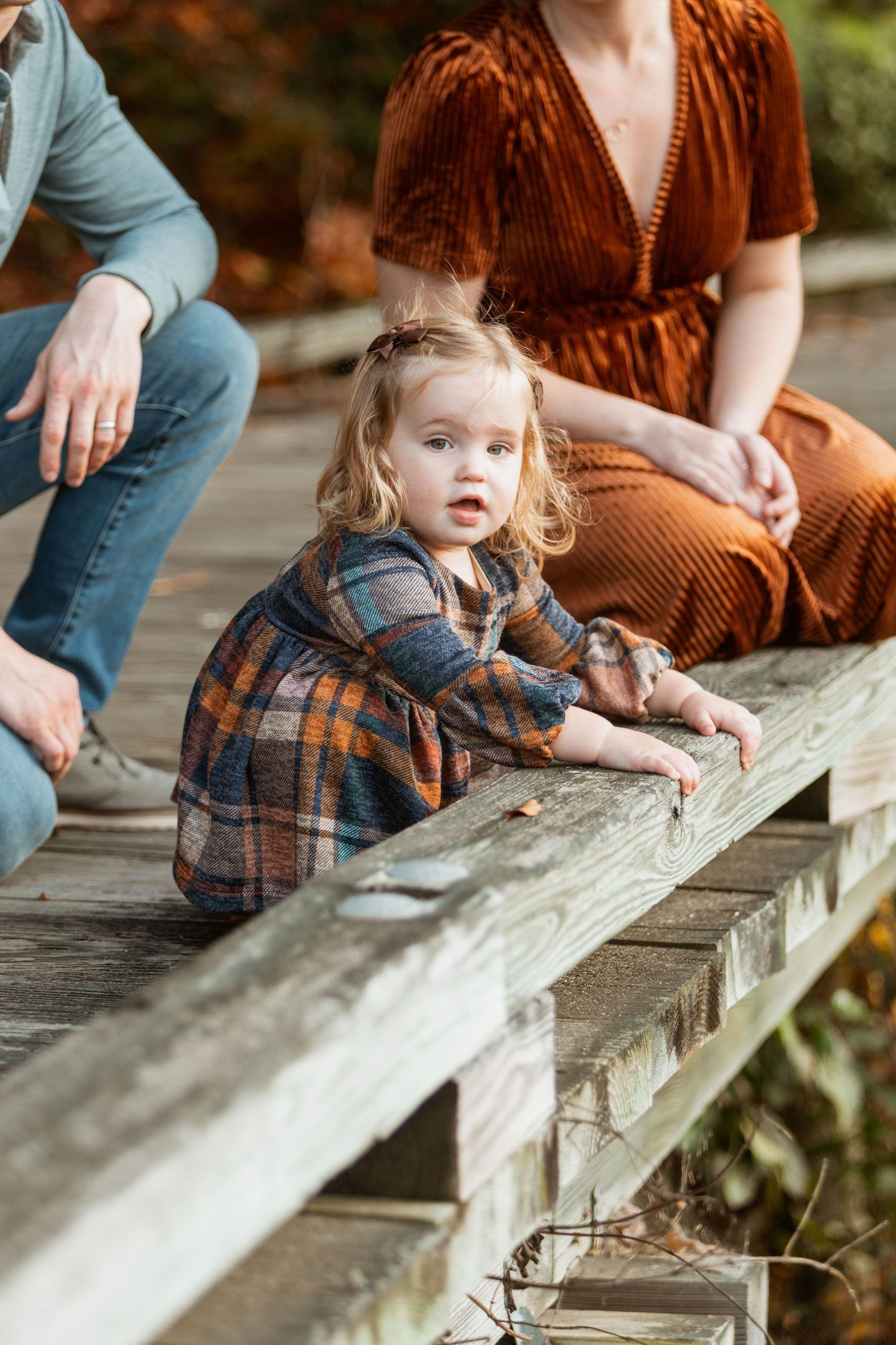 Curious blonde toddler girl in plaid dress leans on wooden boardwalk railing with parents sitting behind her in fall setting.