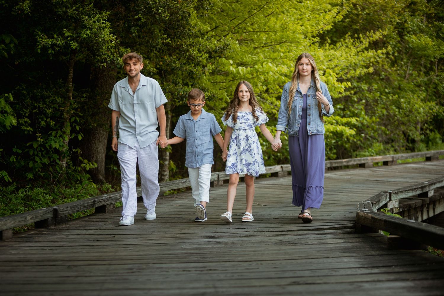 Family of four holding hands walking on a wooden boardwalk surrounded by lush green trees.