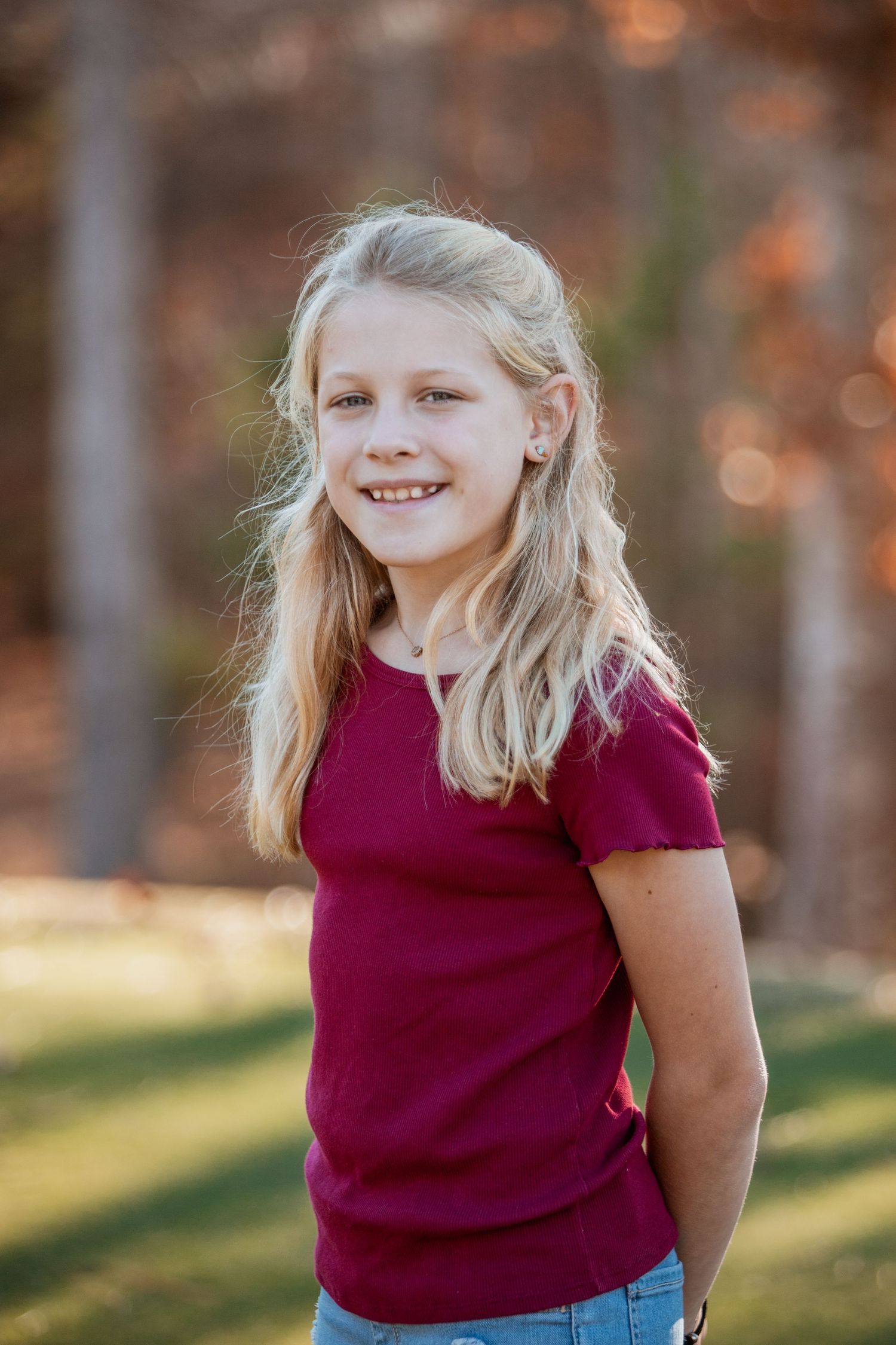 A smiling blonde girl in a burgundy t-shirt stands outdoors with autumn trees in the background.