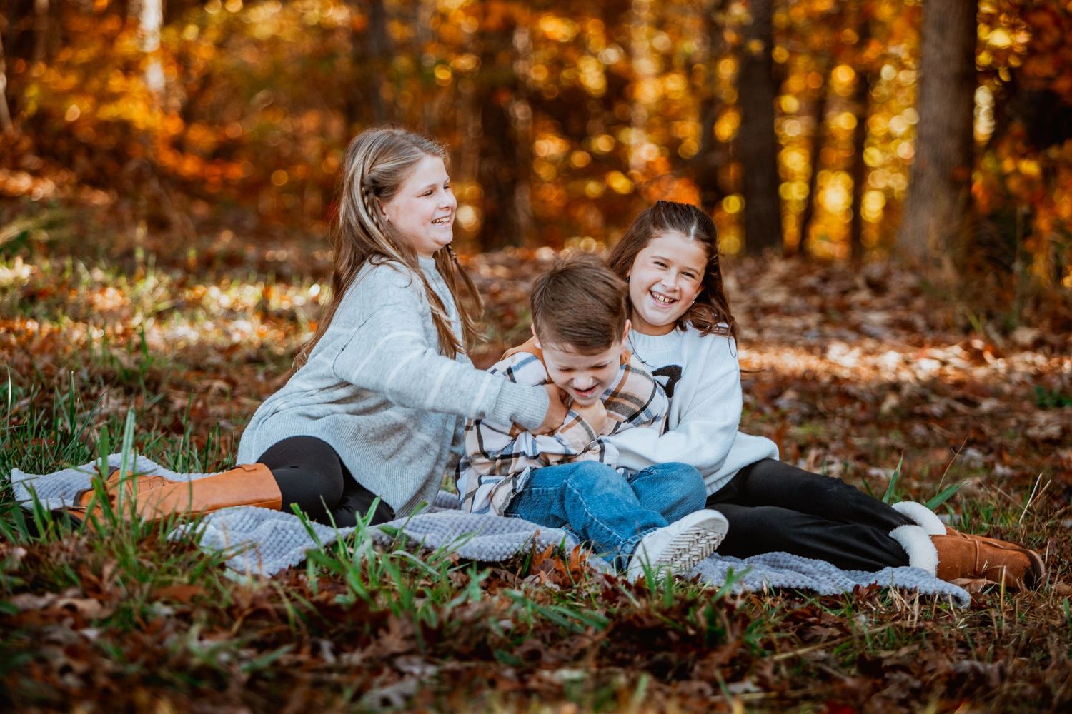 Three children laughing and playing together on a blanket in a colorful autumn forest.