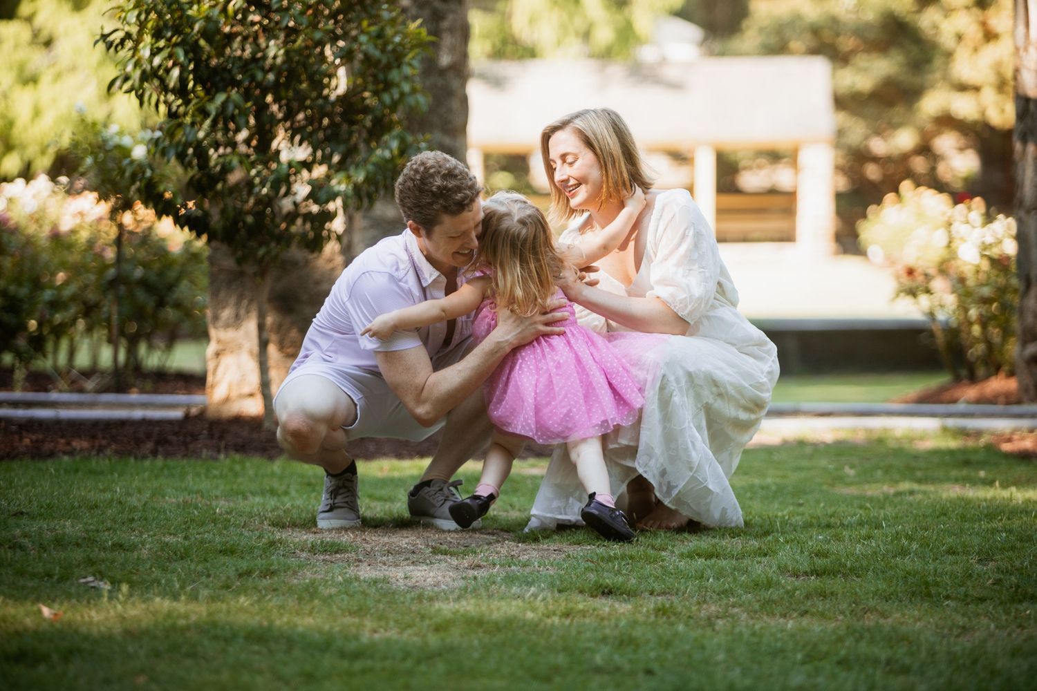 Parents lovingly interact with their young daughter in a pink dress outdoors in a green park setting.