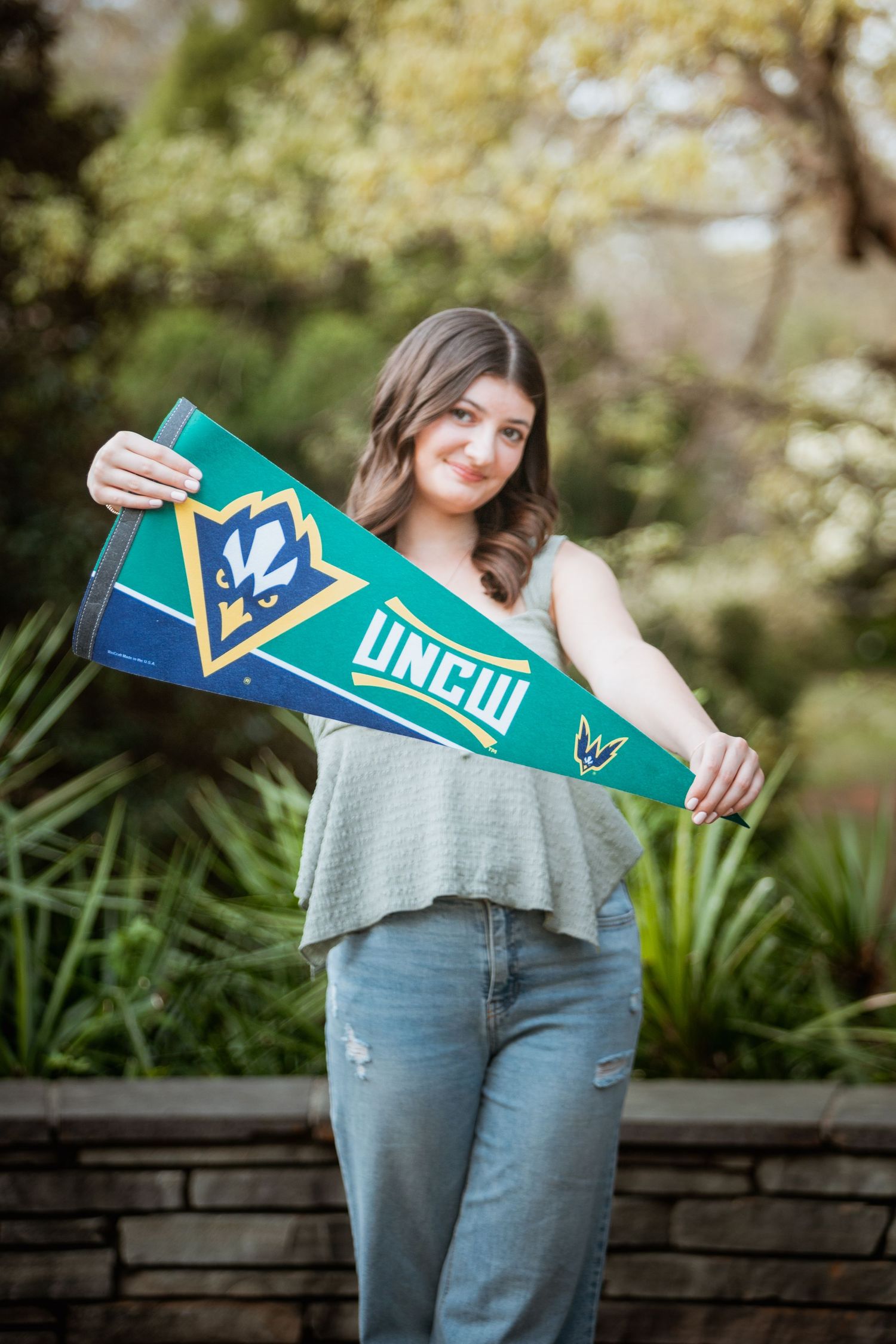 Young woman proudly displaying a UNCW college pennant flag in an outdoor garden.
