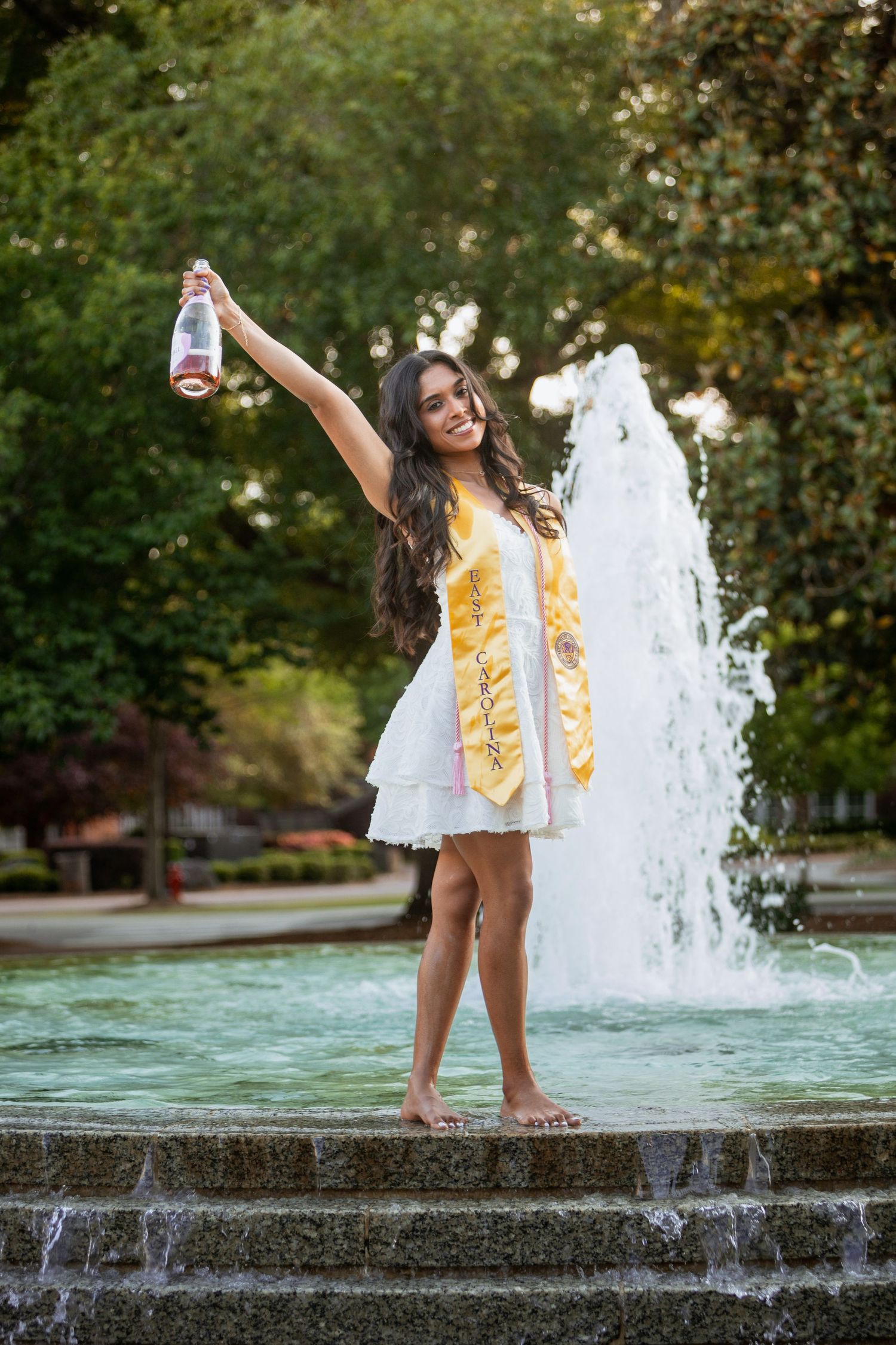 Smiling graduate in white dress and gold stole celebrating barefoot at campus fountain holding bottle.