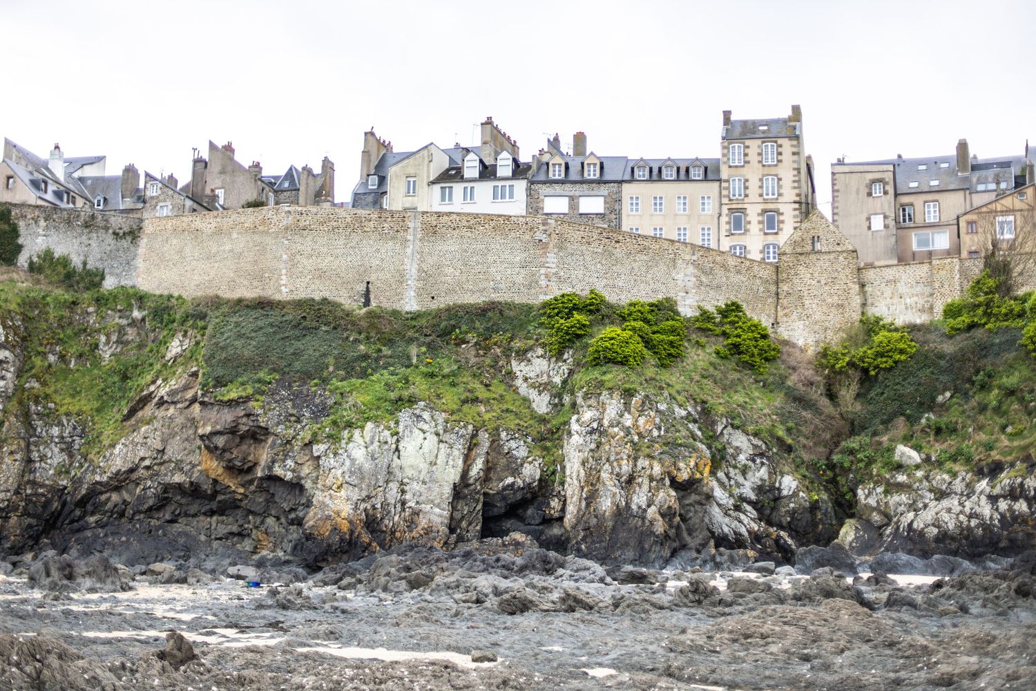 Ancient stone city walls and historic buildings perched on rocky coastal cliffs under an overcast sky.