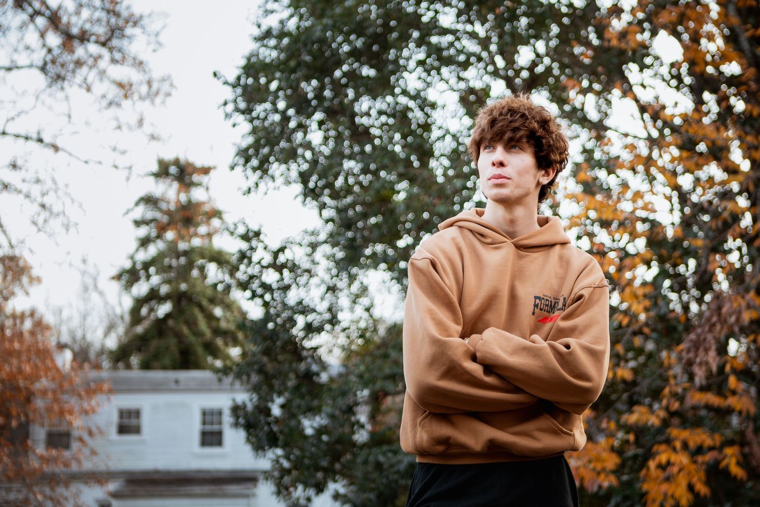 Young man with curly hair in a tan hoodie standing with arms crossed among autumn trees.