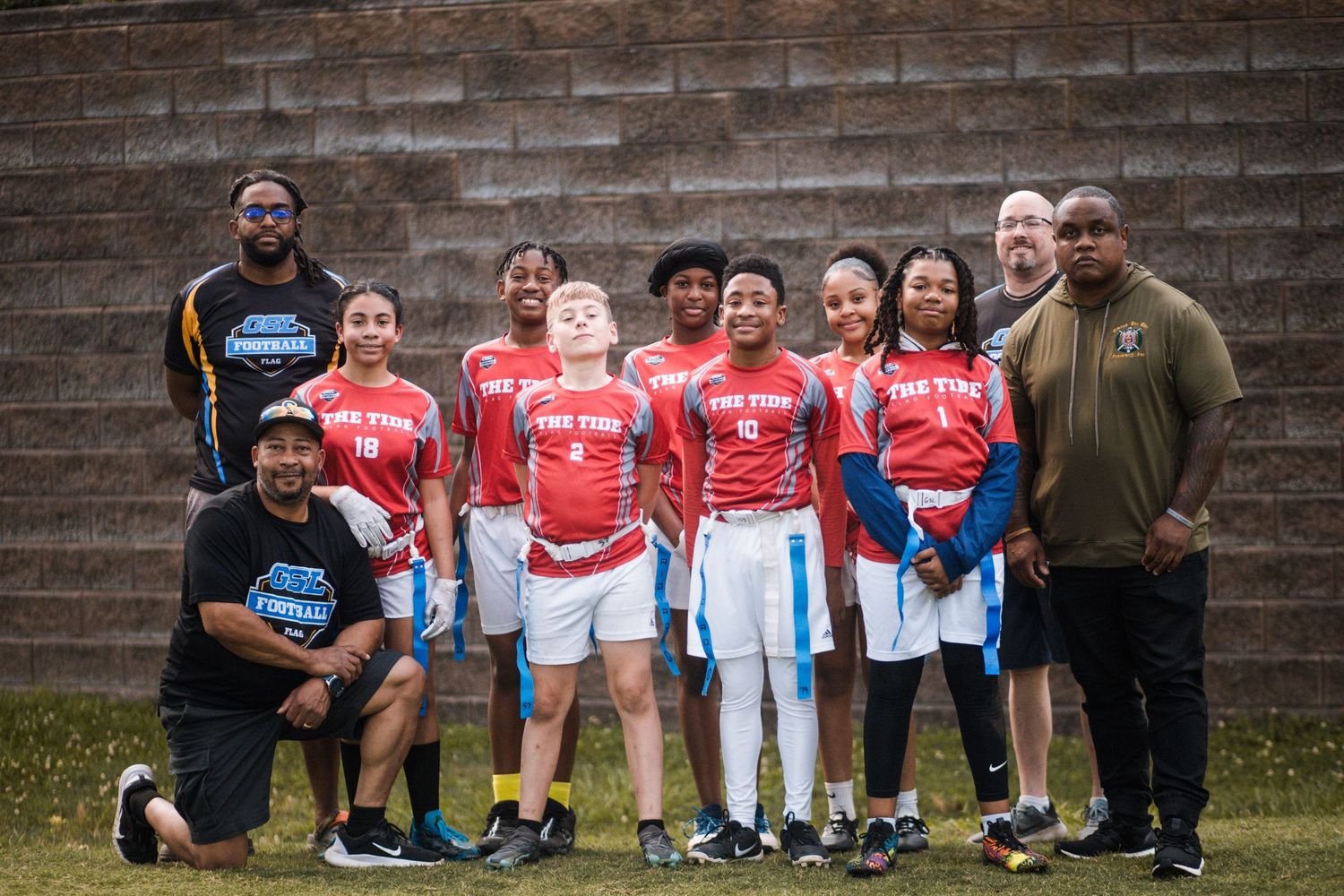 Youth flag football team in red Fire uniforms posing with coaches against a brick wall.