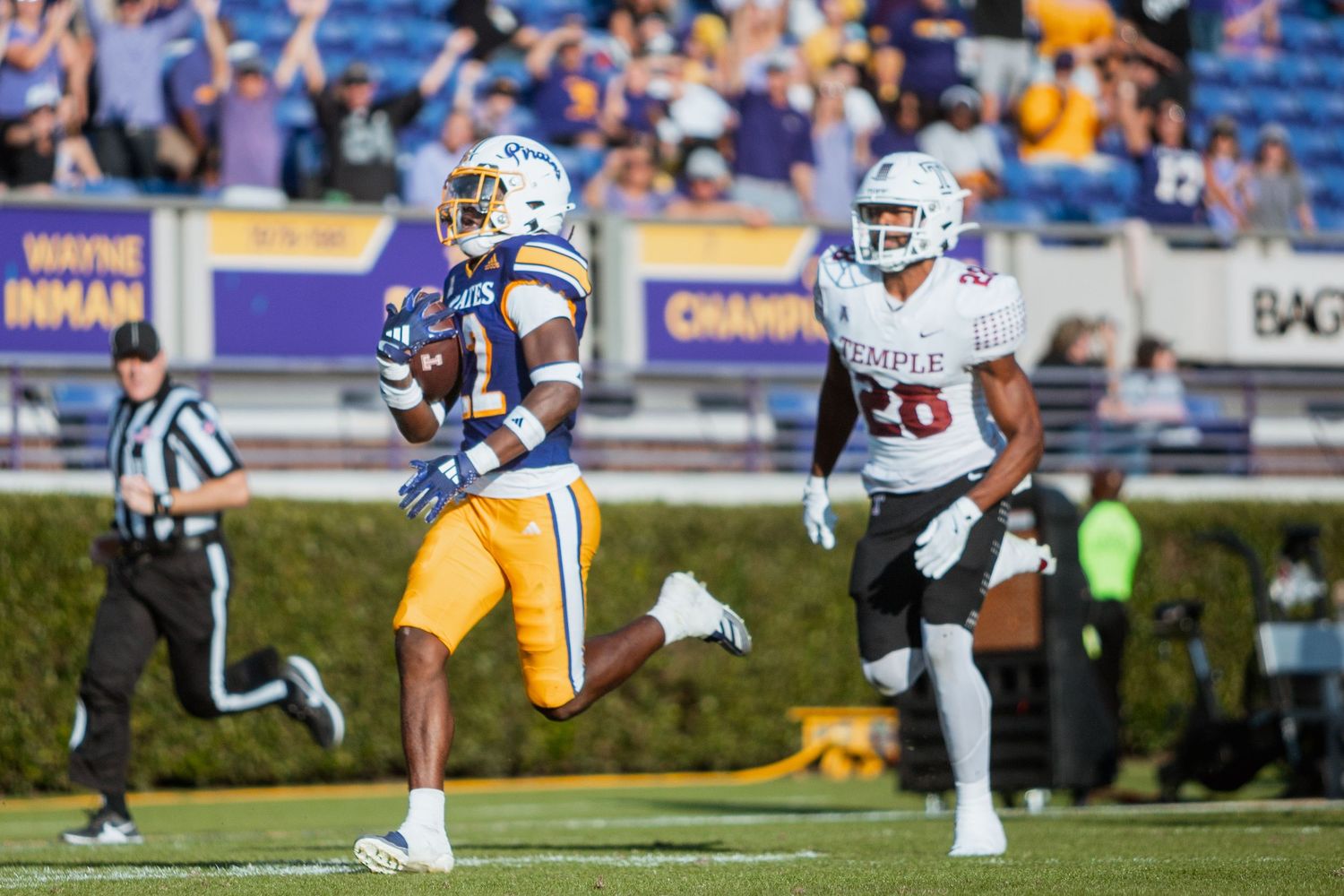 ECU Pirates player scores a touchdown while a Temple defender trails behind at Dowdy-Ficklen Stadium.