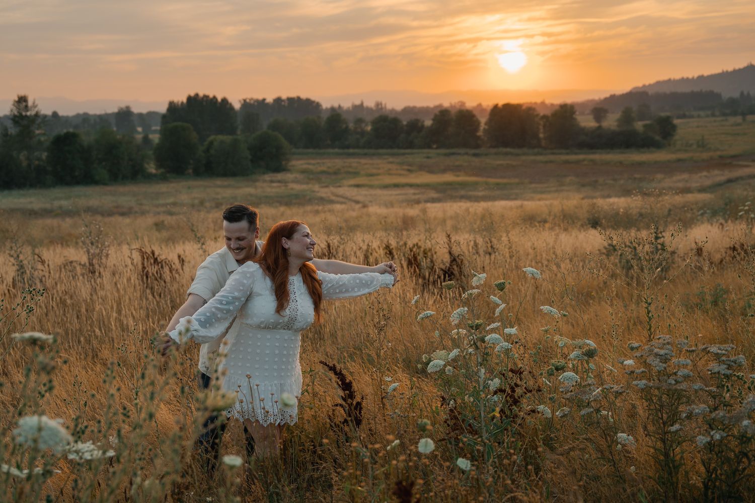 Couple embracing in golden field at sunset, woman in white lace dress with red hair, romantic outdoor photography.