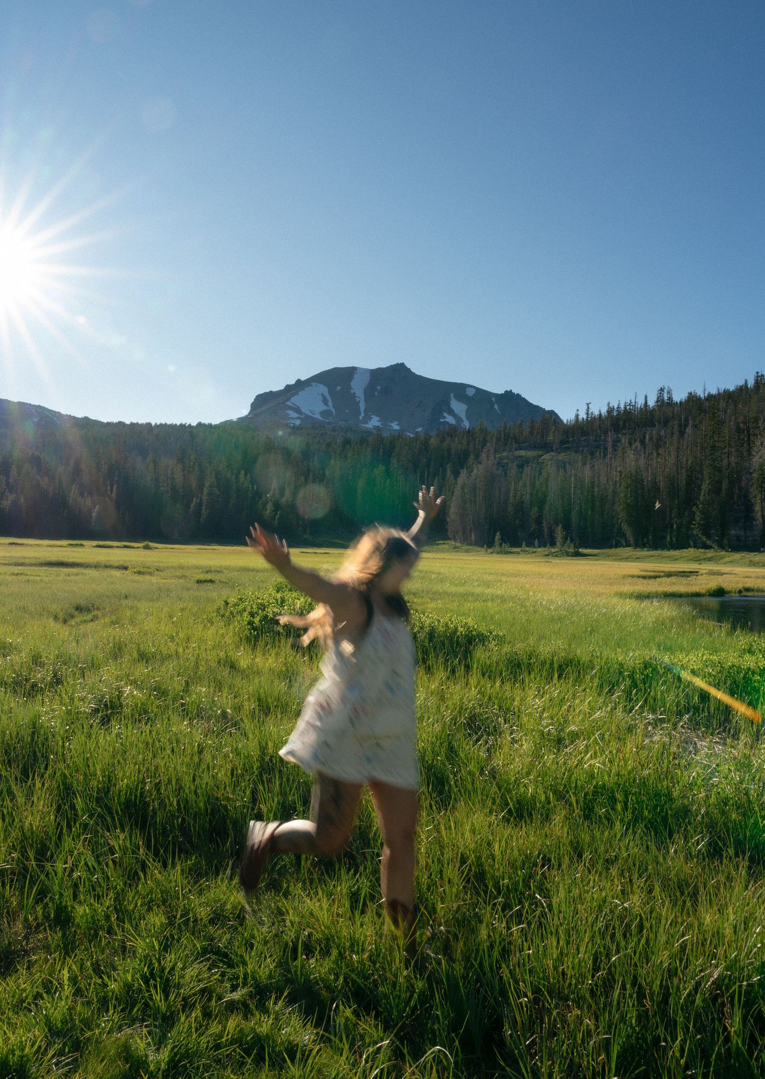 Young girl twirling in a green meadow with a mountain backdrop and bright sun on a clear day.