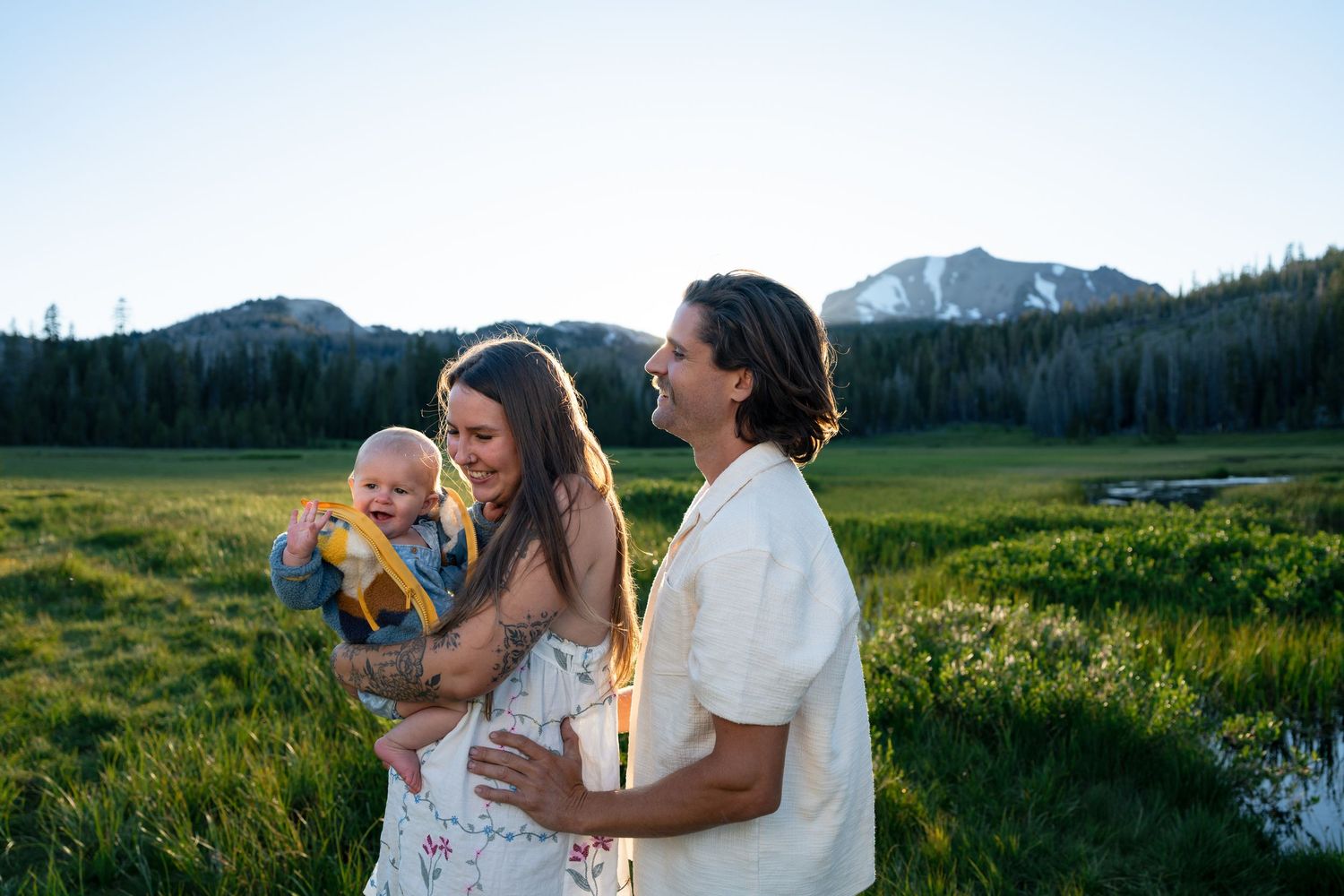 Happy family with baby enjoying golden hour in a lush mountain meadow with scenic peaks and forest backdrop.