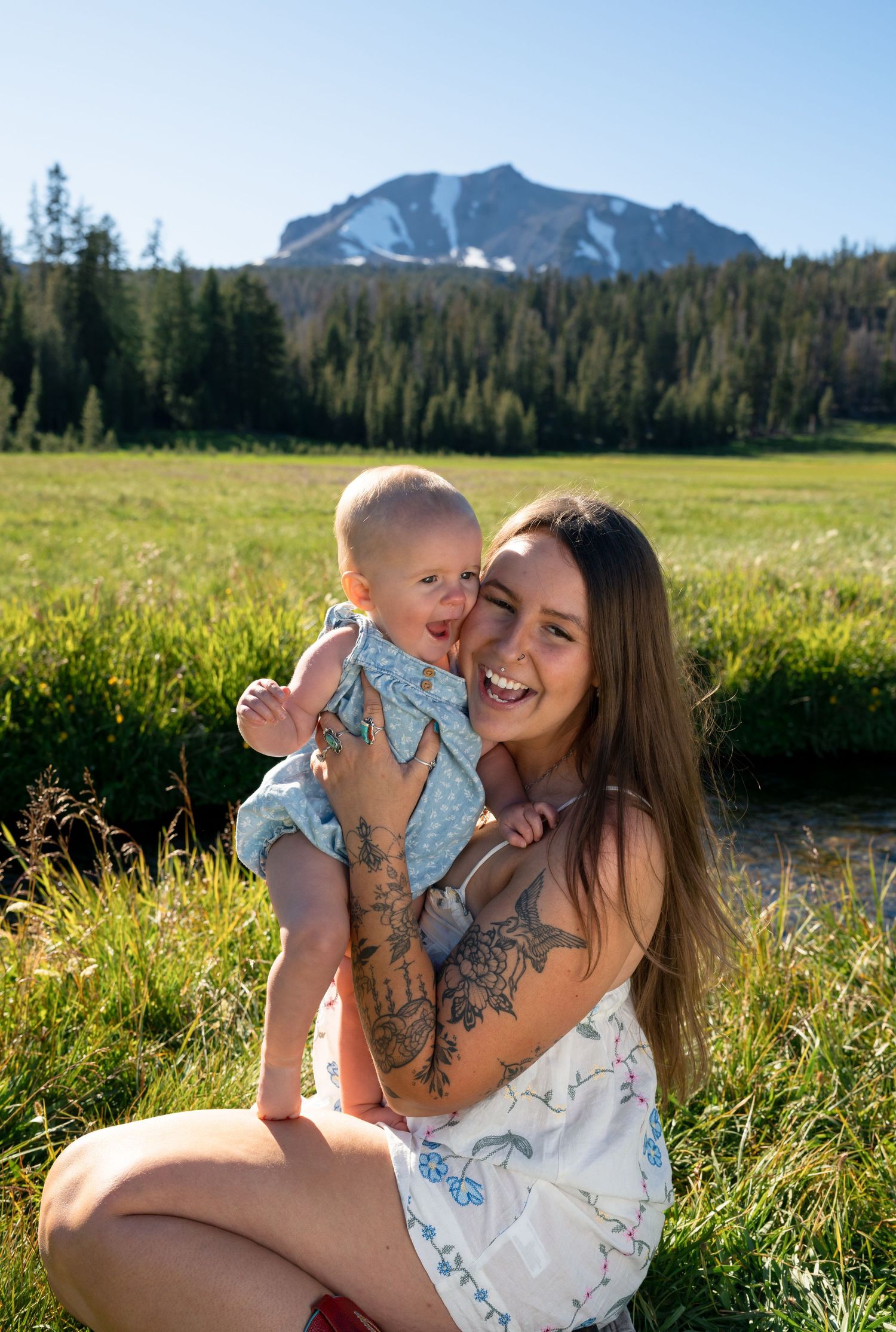 Smiling young mother with tattoos holds laughing baby in sunny mountain meadow with pine trees and snowy peak behind.