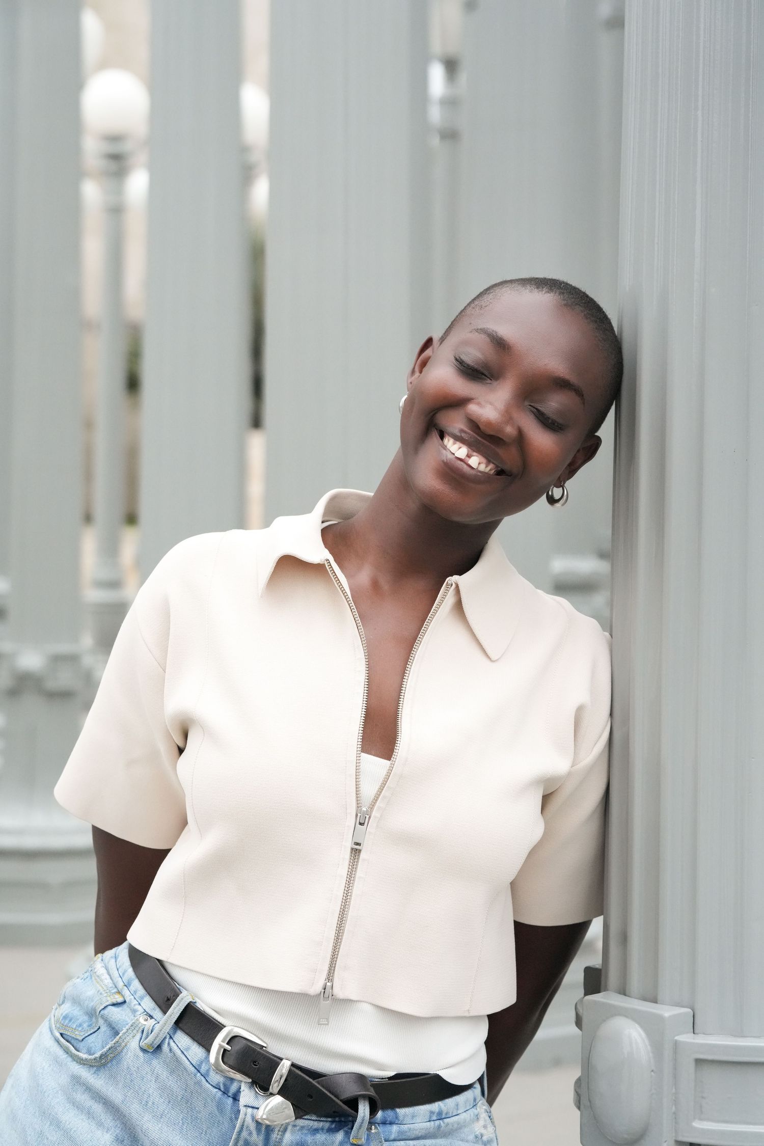 Smiling woman in cream zip-up top and jeans leaning against gray column outdoors.