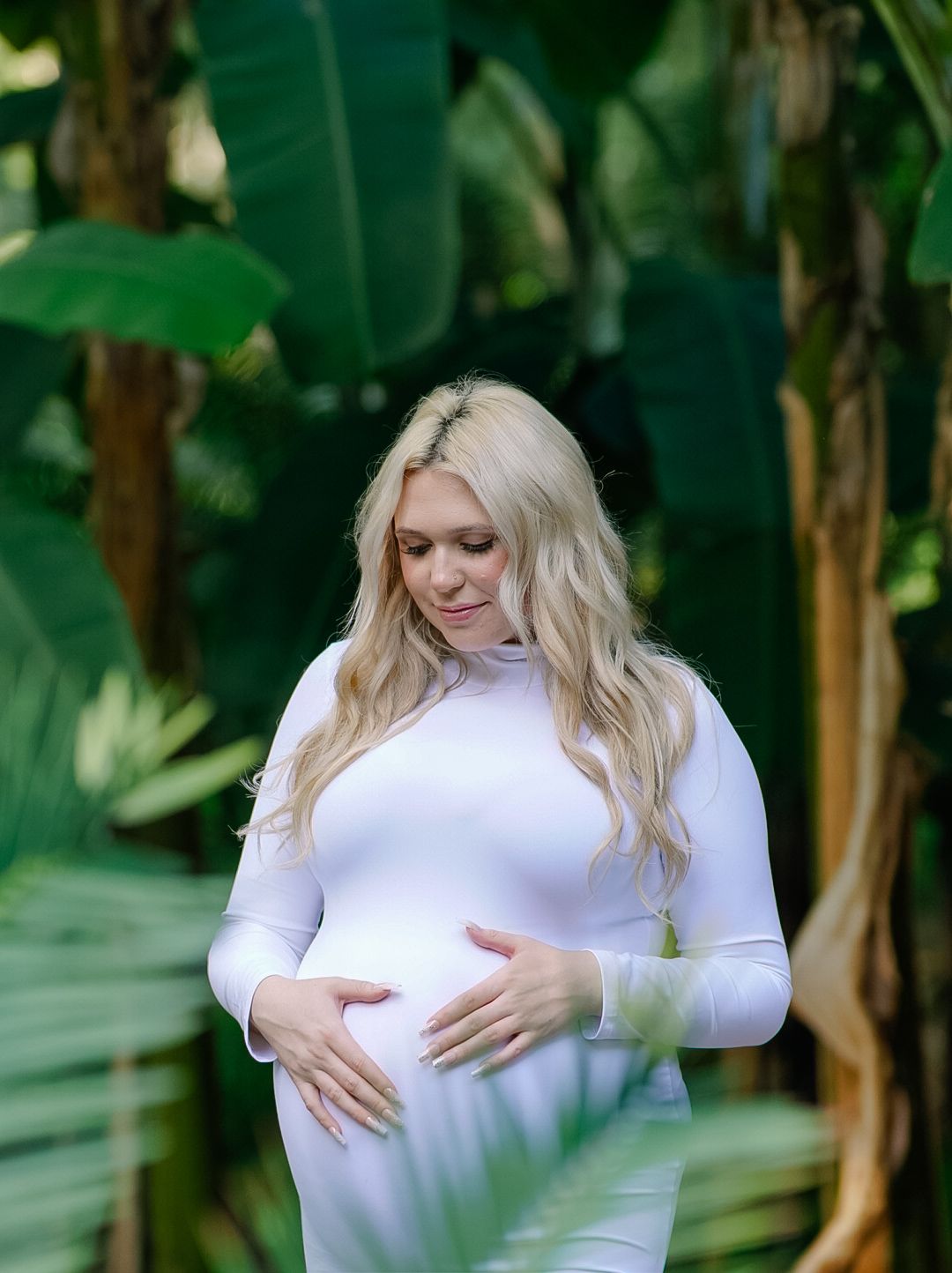 Pregnant woman in white dress cradling baby bump outdoors surrounded by lush tropical green leaves.