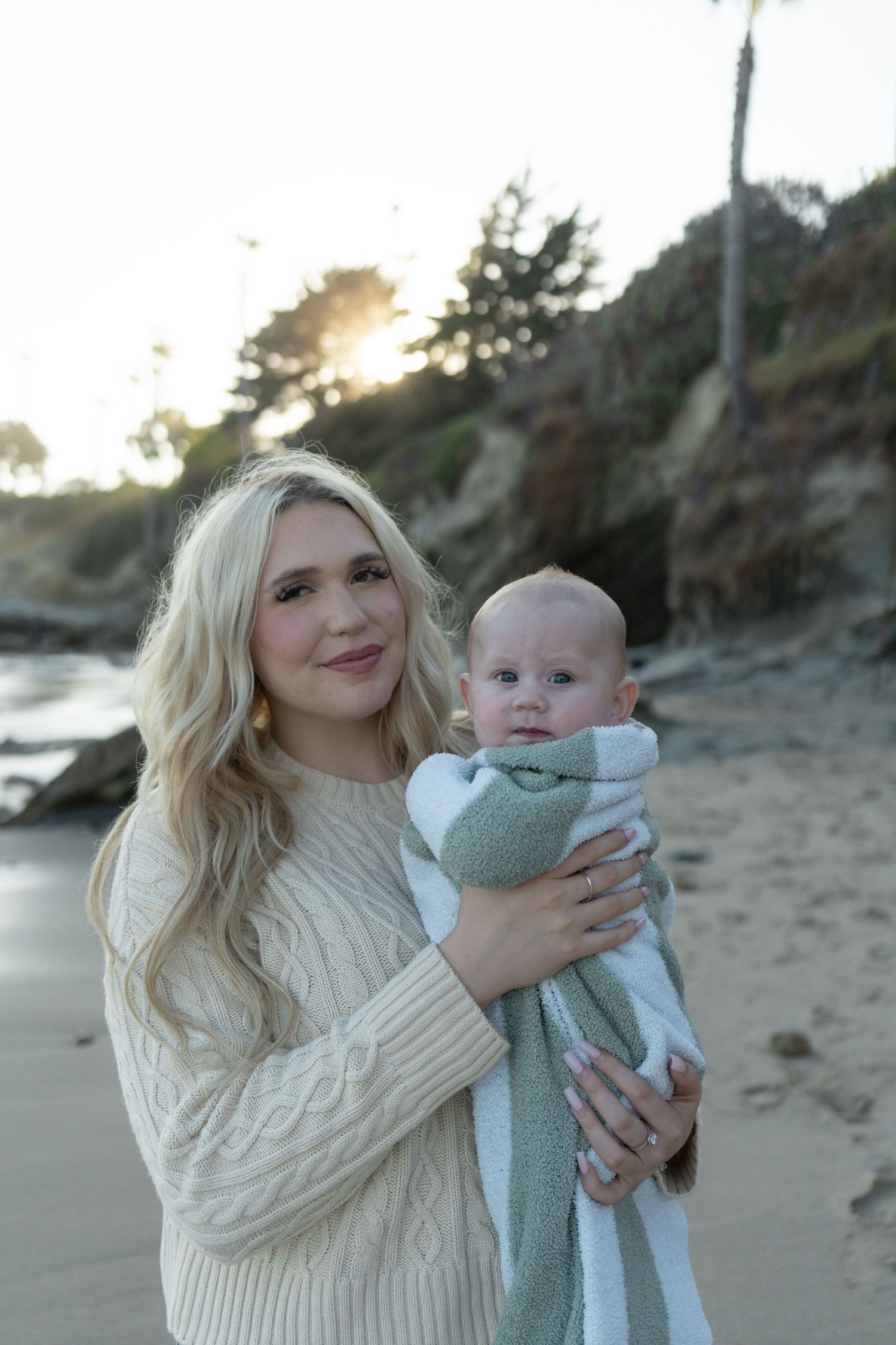Blonde woman in cream cable-knit sweater holds a swaddled baby on a sandy beach at sunset.