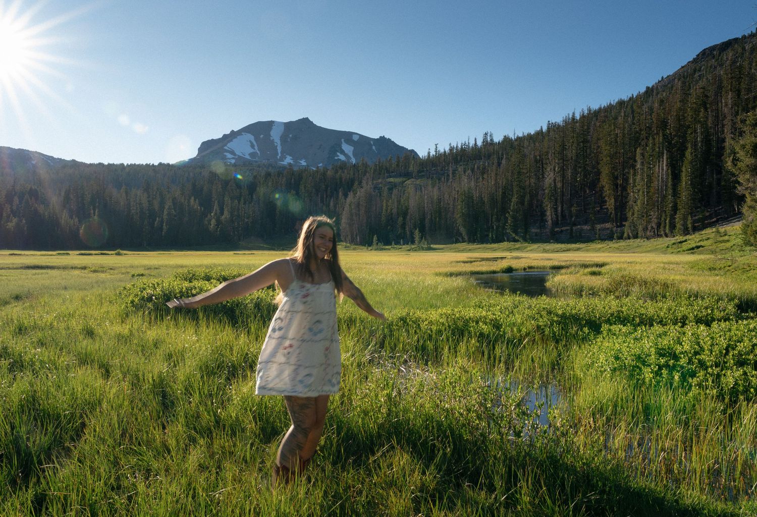 Woman in white dress standing in lush green mountain meadow with snow-capped peak and forest backdrop on sunny day.
