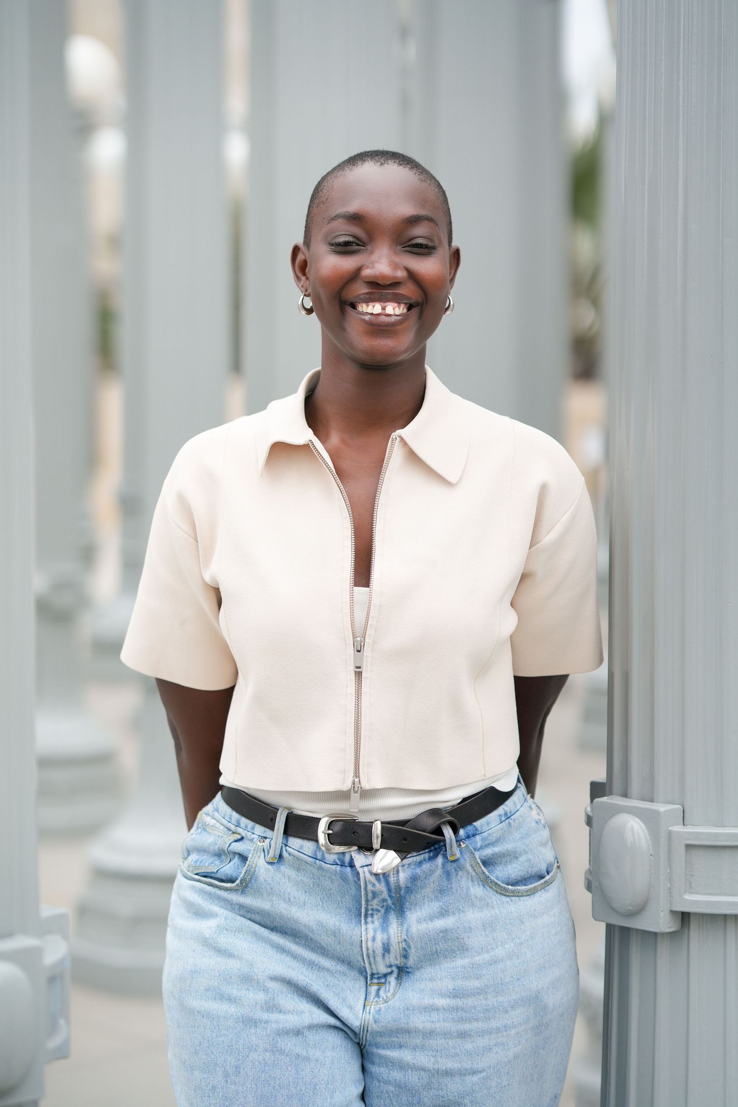 Smiling young Black woman with shaved head wearing beige zip shirt and jeans, posing outdoors near gray columns.