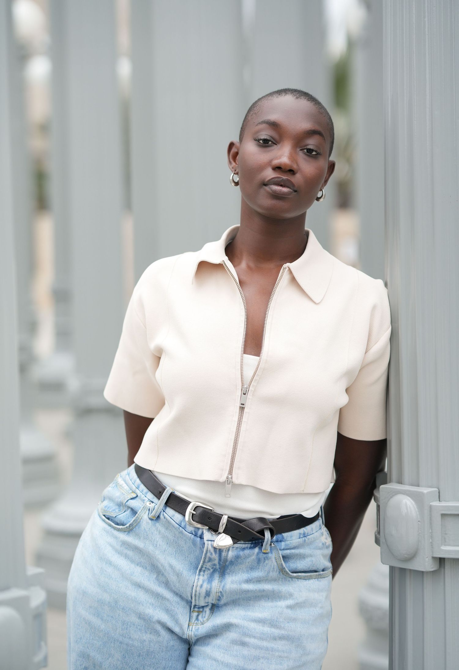 A young Black woman with a shaved head wearing a cream zip-up shirt and light blue jeans leans against a gray pillar.