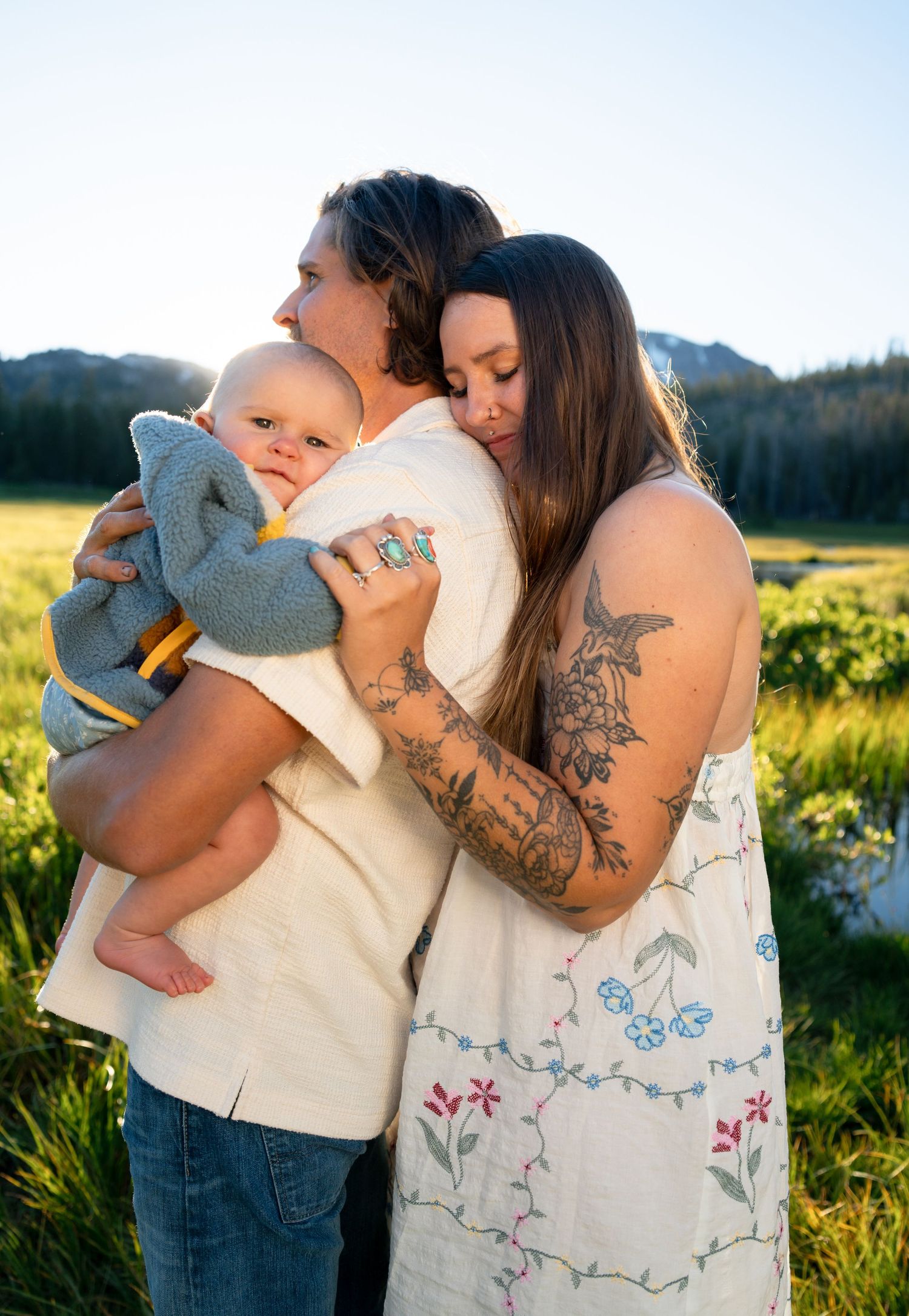 A couple embraces outdoors in a meadow, holding their baby wrapped in a gray blanket, with mountains in the background.