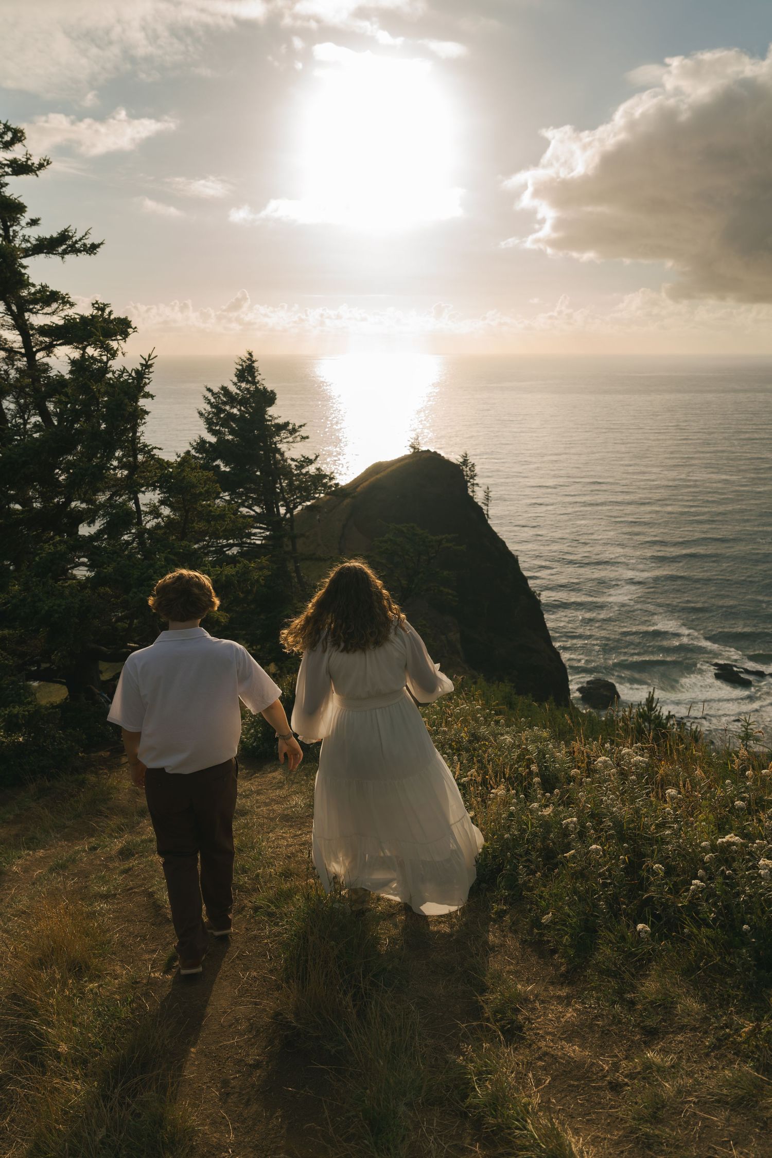 Couple holding hands walking along coastal cliff path at sunset, woman in white dress, ocean and rocky headland in background.
