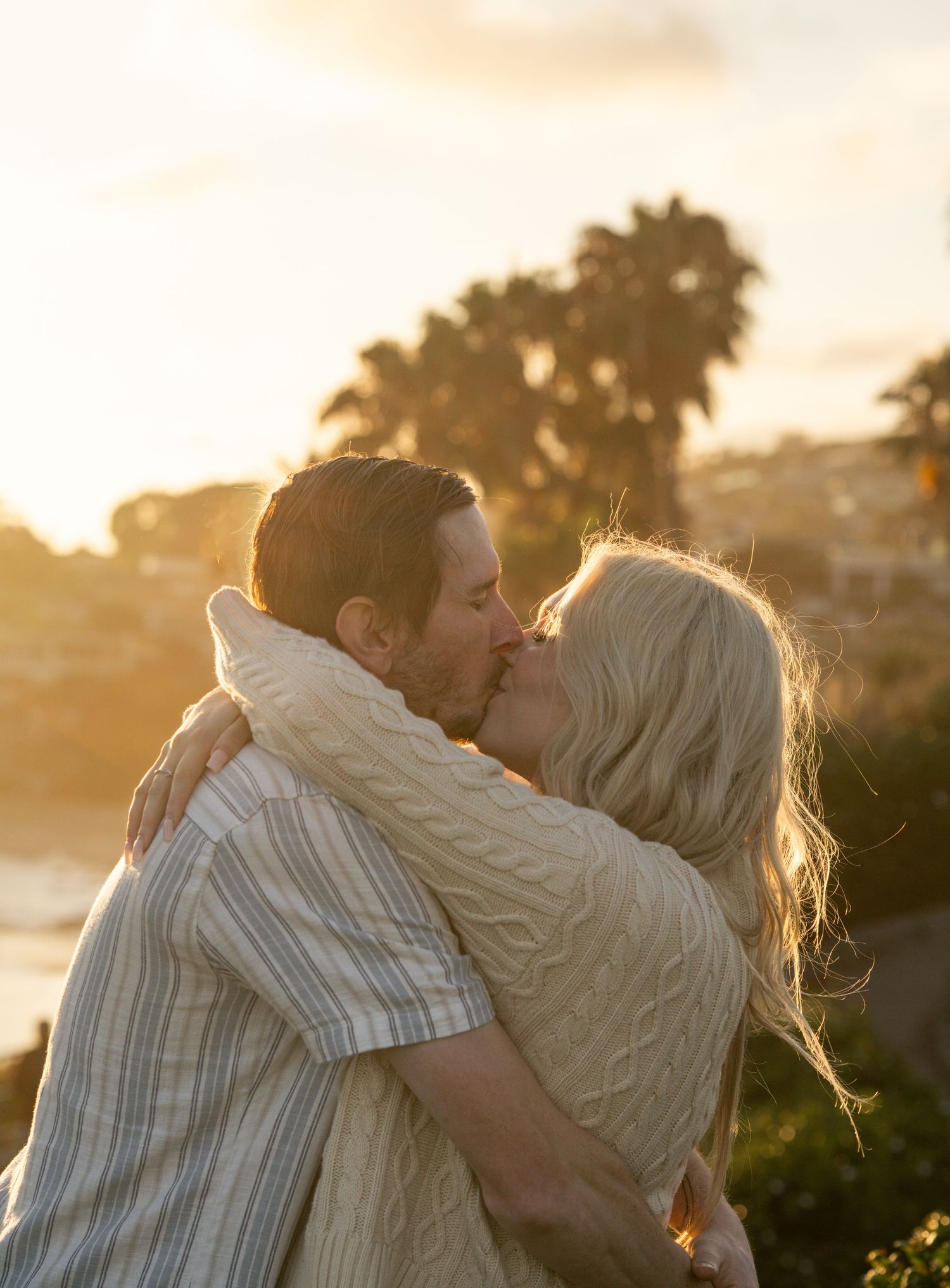 Couple sharing a romantic kiss at golden hour sunset with palm trees and ocean view in background.