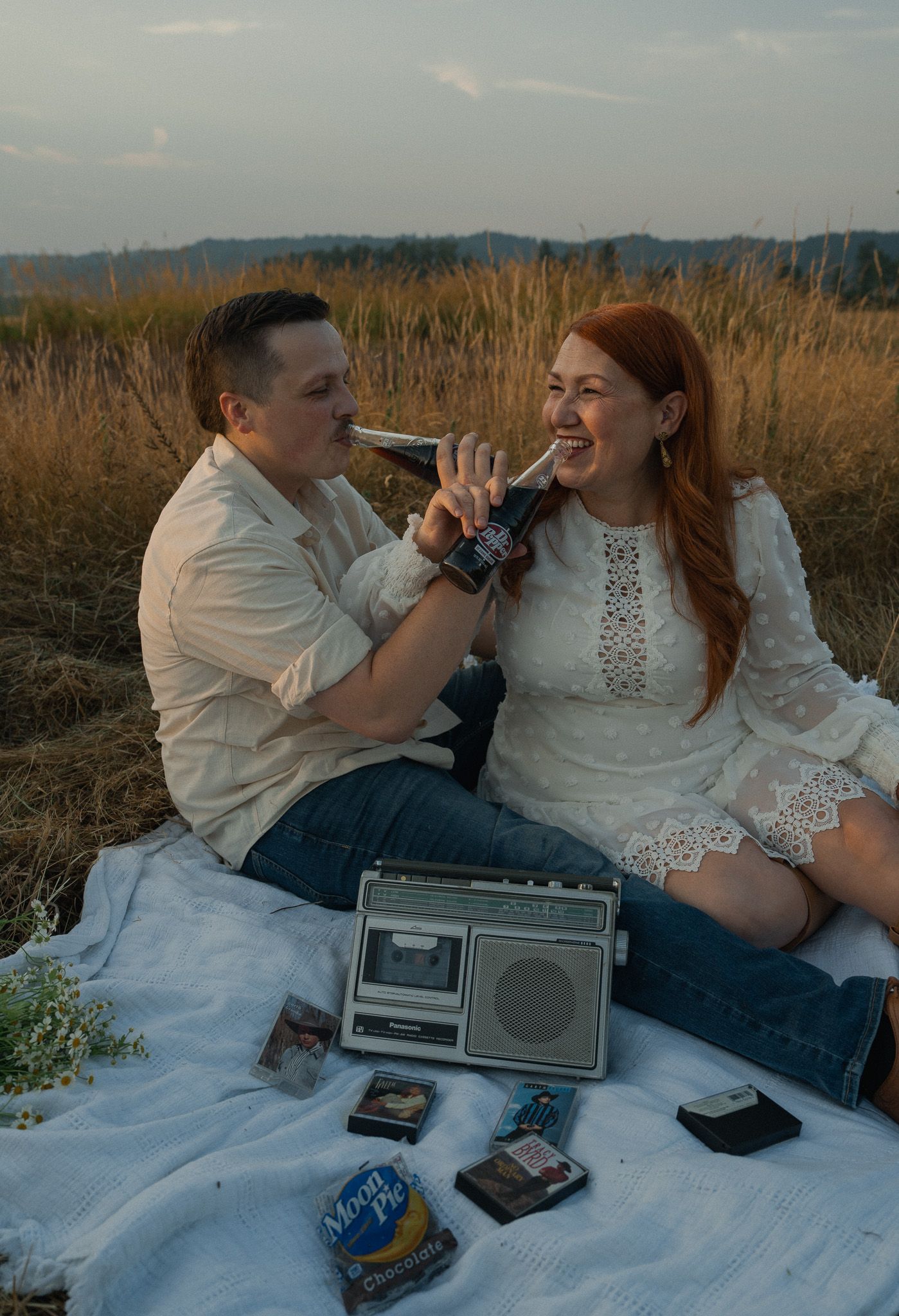 Couple sharing a Dr. Pepper bottle during a romantic picnic in a golden field with a vintage cassette player nearby.