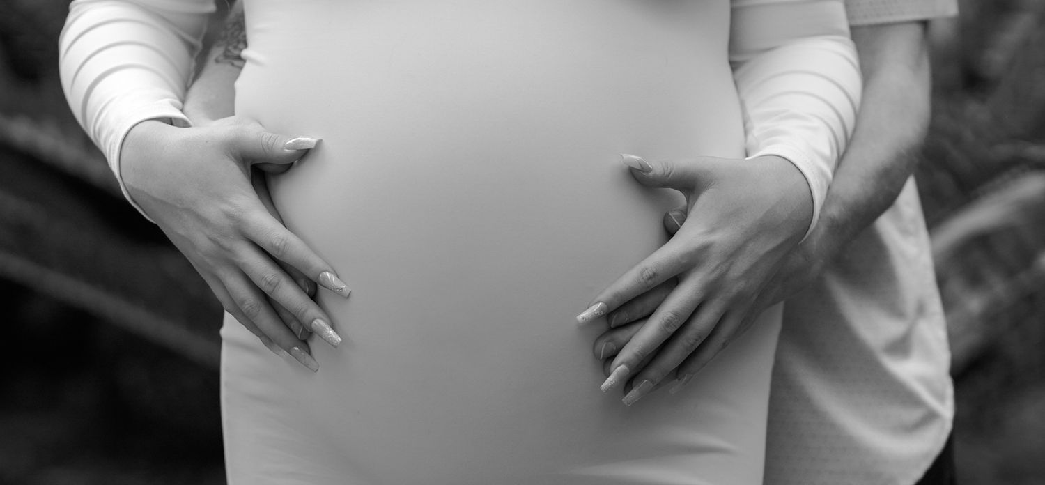 Black and white photo of a pregnant woman cradling her baby bump with both hands outdoors.