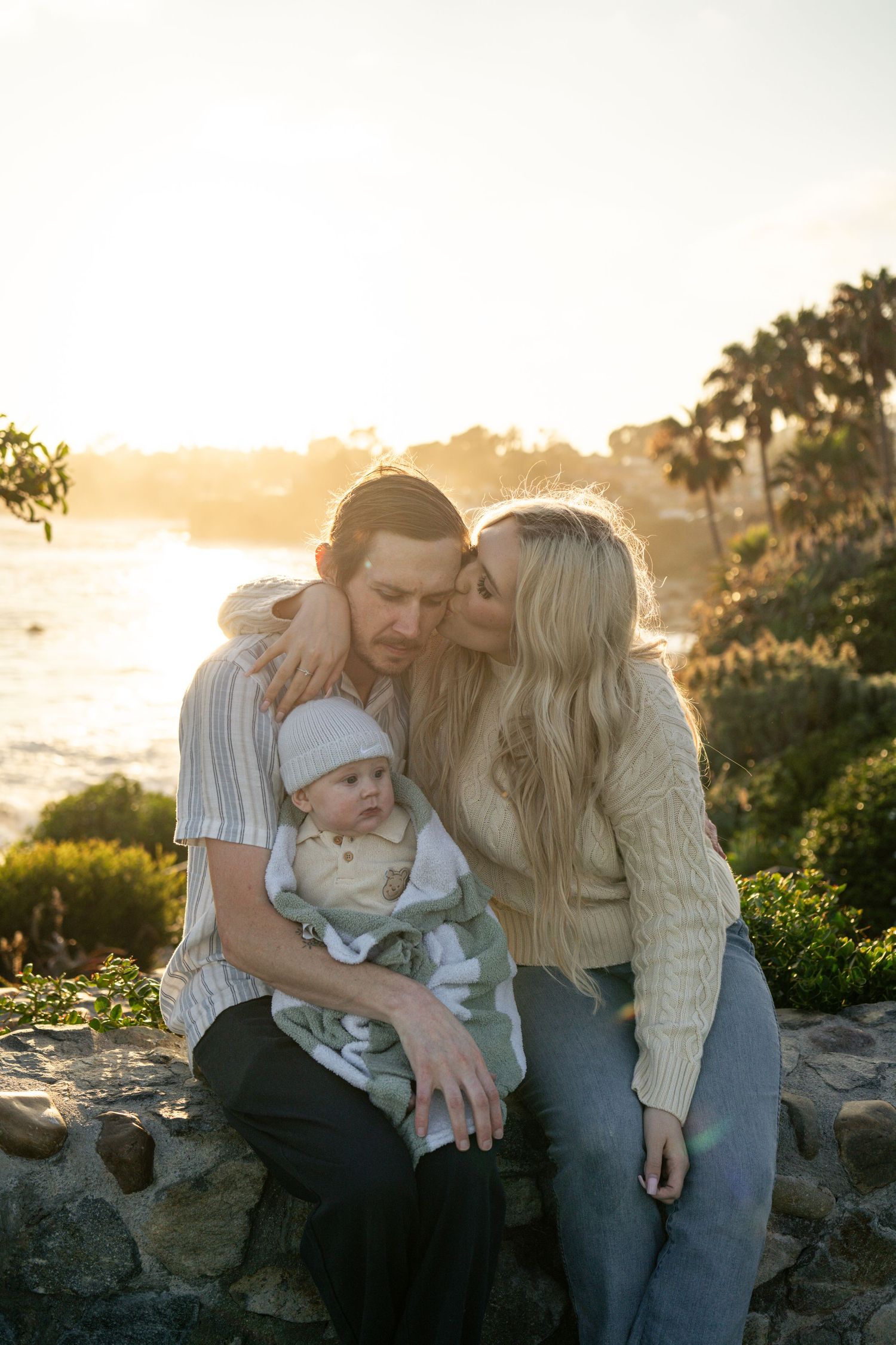 A couple shares a tender moment with their baby at sunset near the ocean, surrounded by lush greenery and palm trees.