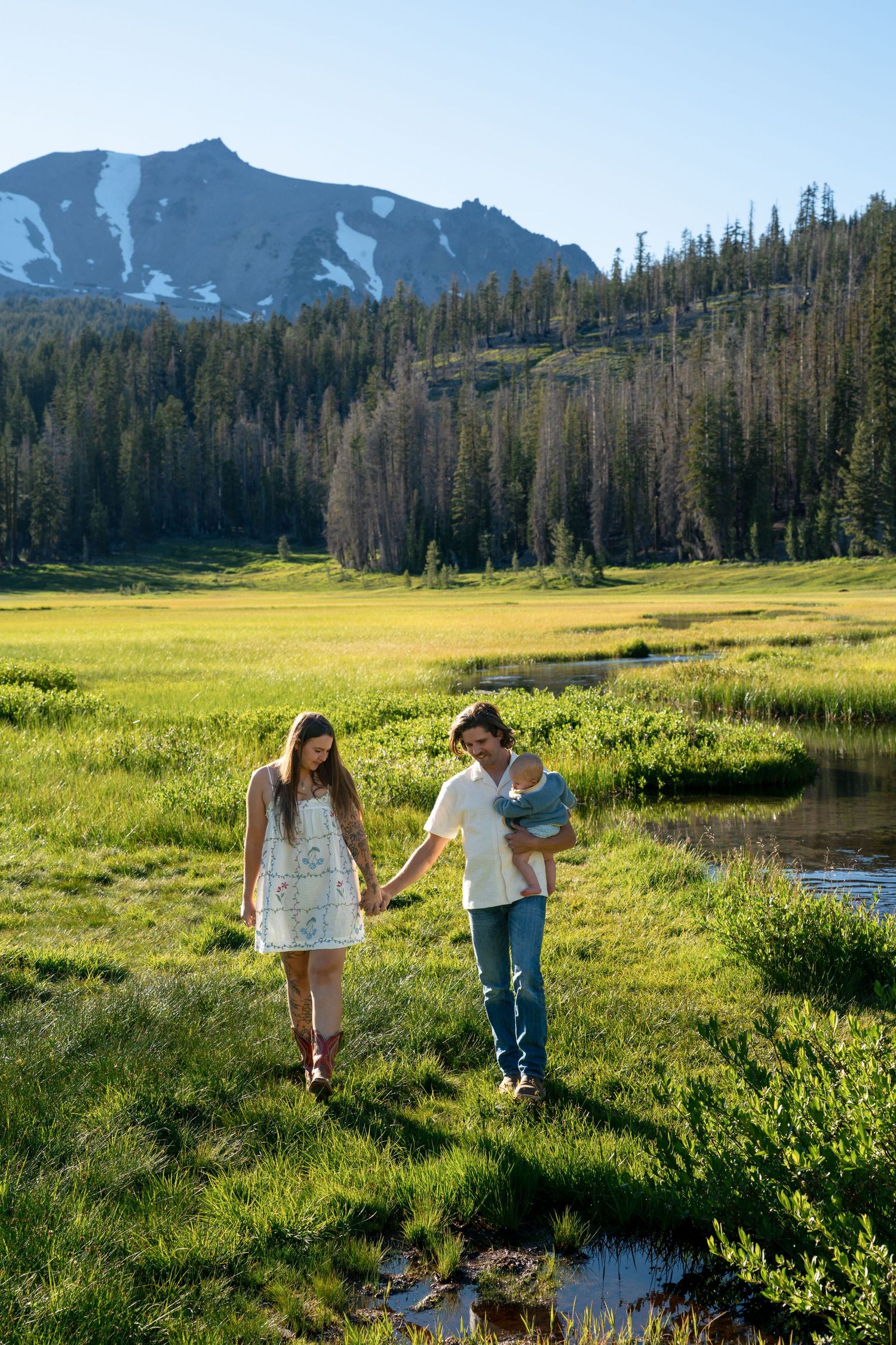 A young family walks through a lush green mountain meadow, with a snow-capped peak and pine forest in the background.