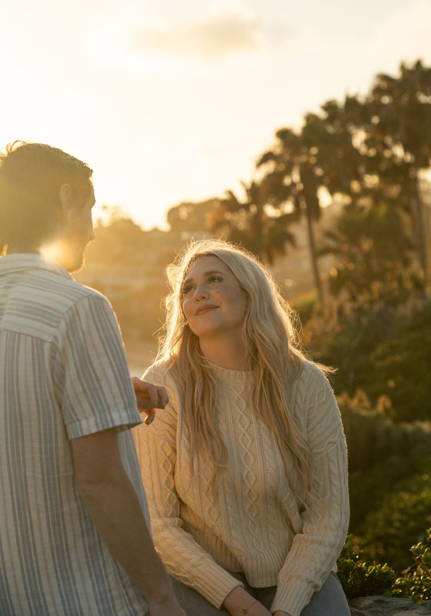 A young blonde woman in a cream cable-knit sweater gazes up at a man during a golden sunset outdoors.