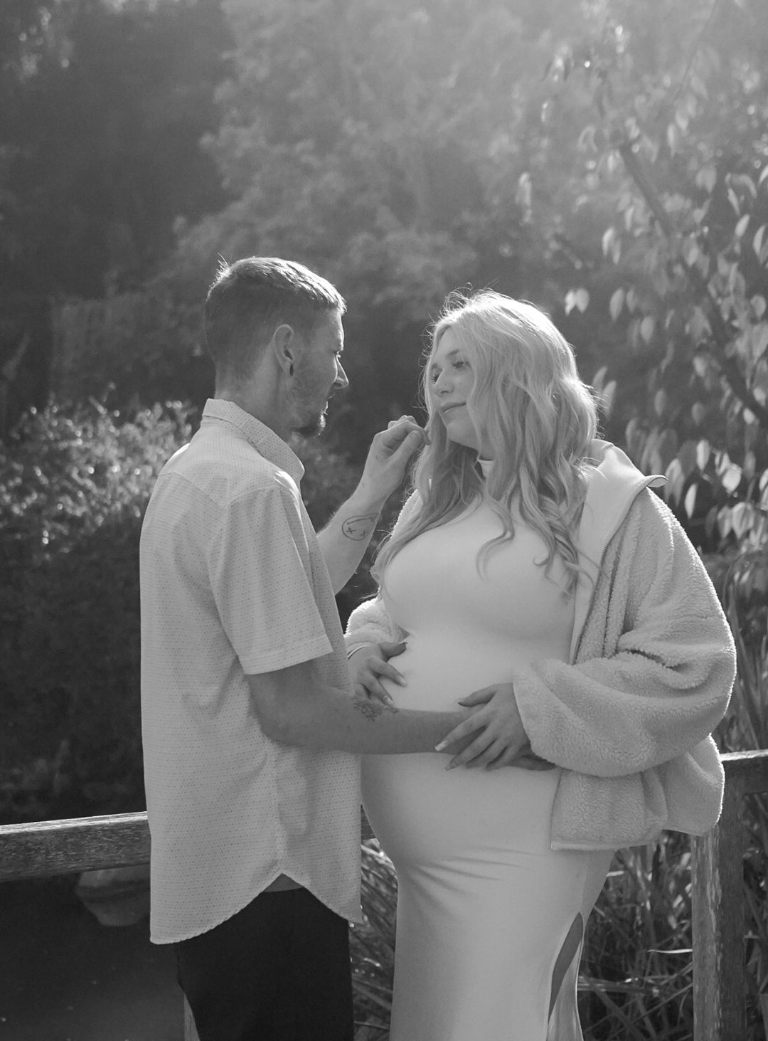 Black and white maternity photo of a couple outdoors, man tenderly touching womans face as she cradles her baby bump.