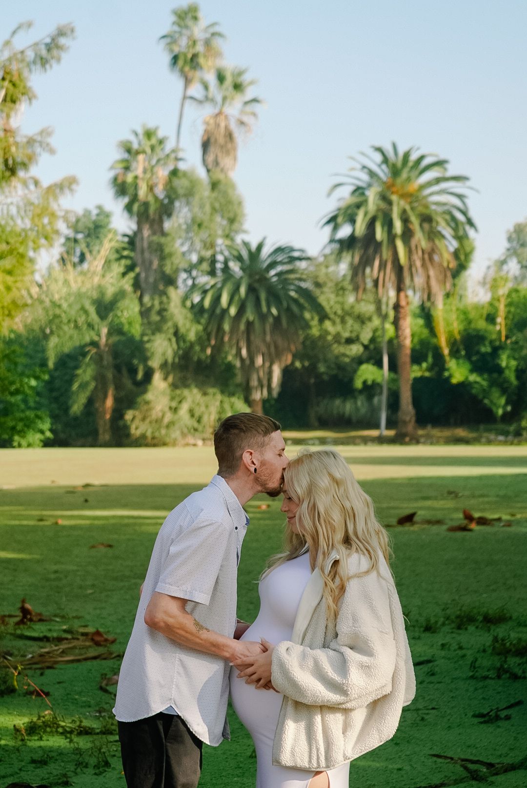 Couple sharing a kiss outdoors during a maternity photoshoot with lush palm trees in the background.