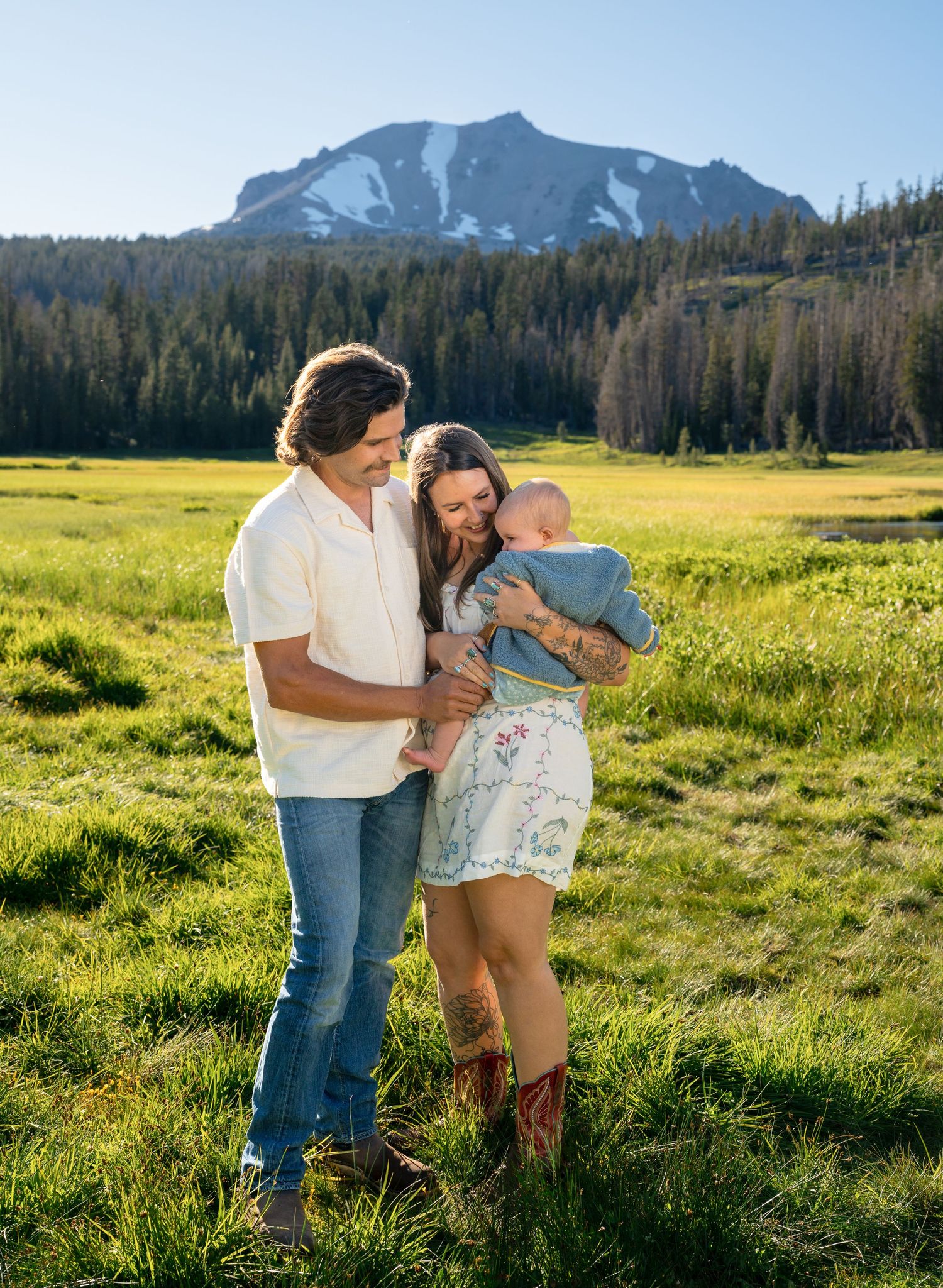 Young family with newborn baby posing in a green meadow with snow-capped mountain and pine forest backdrop.