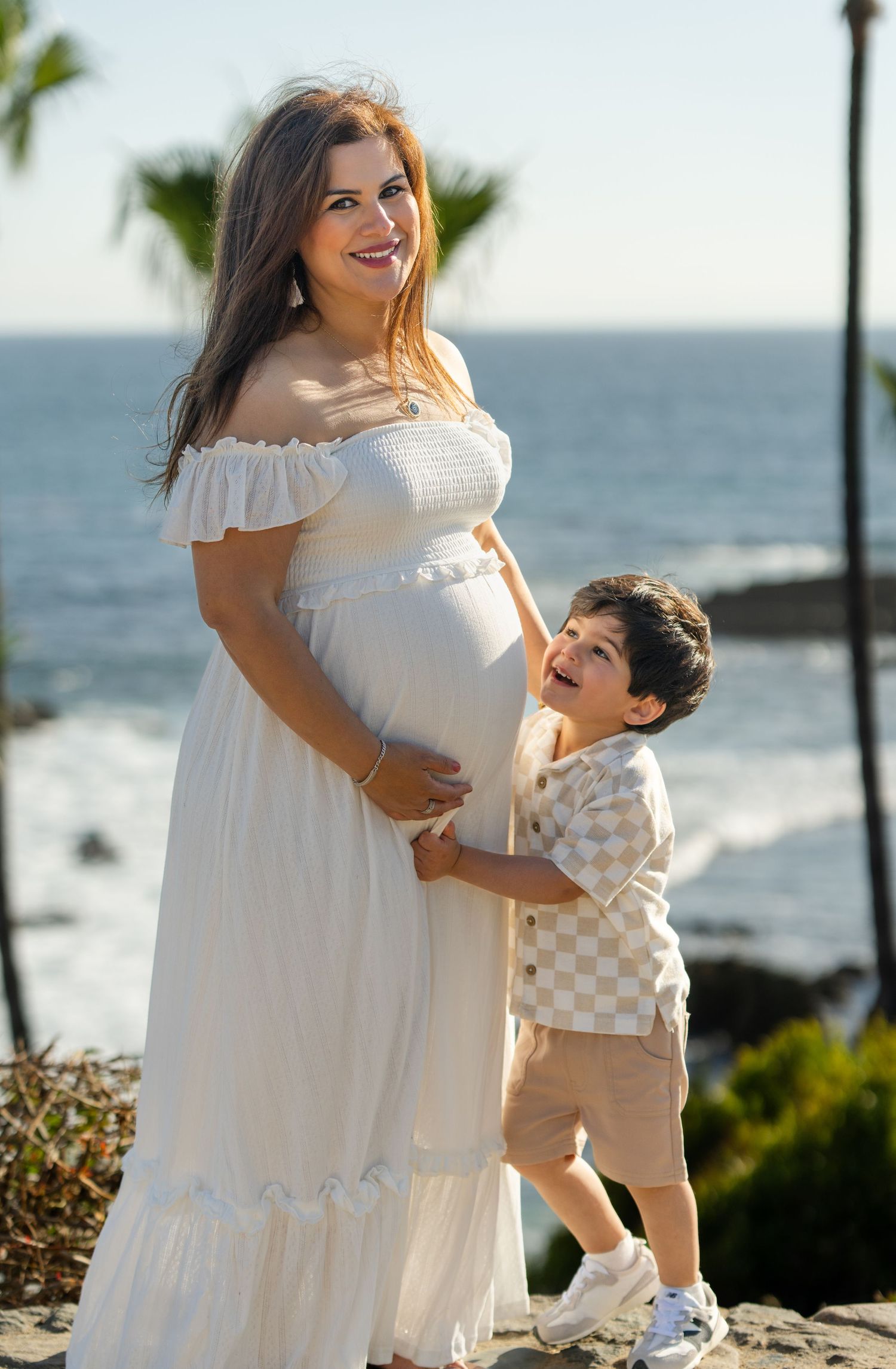 Pregnant woman in white off-shoulder dress poses with young boy hugging her belly near ocean.