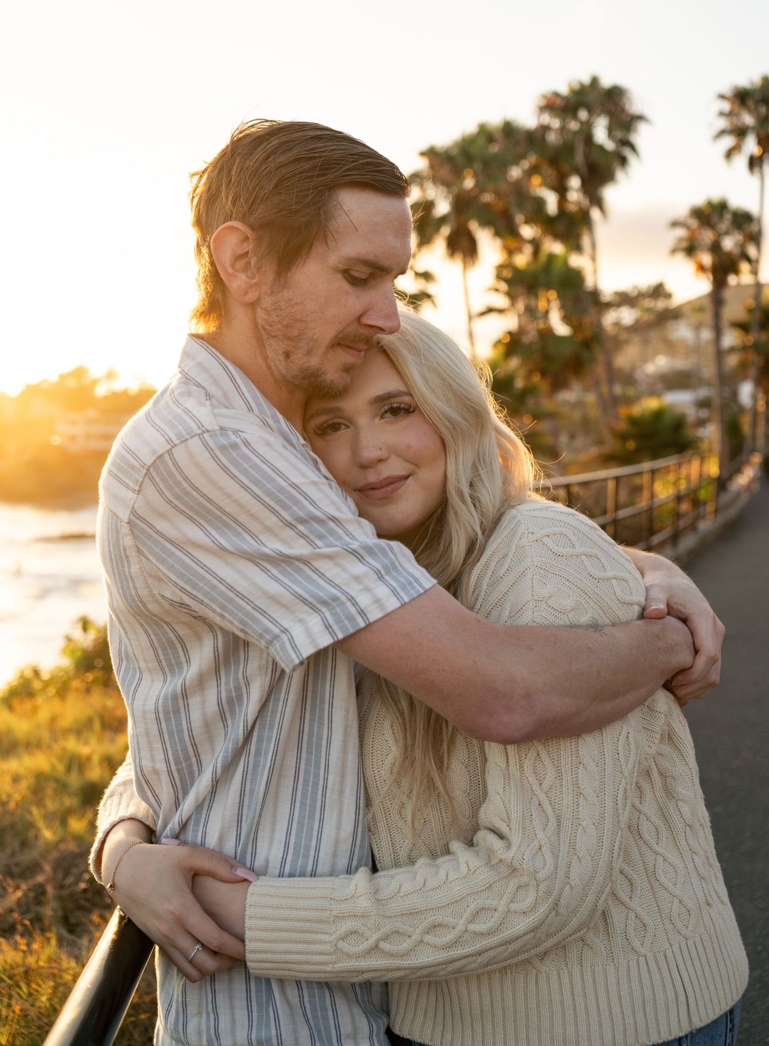 Couple embracing at golden hour near coastal palm trees, woman in cable-knit sweater smiling warmly.