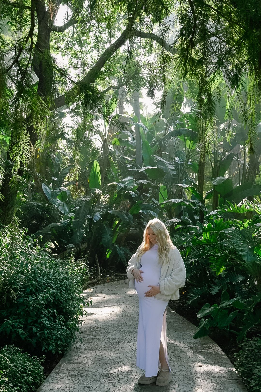 Pregnant woman in white dress and cream cardigan standing on garden path surrounded by lush tropical greenery.