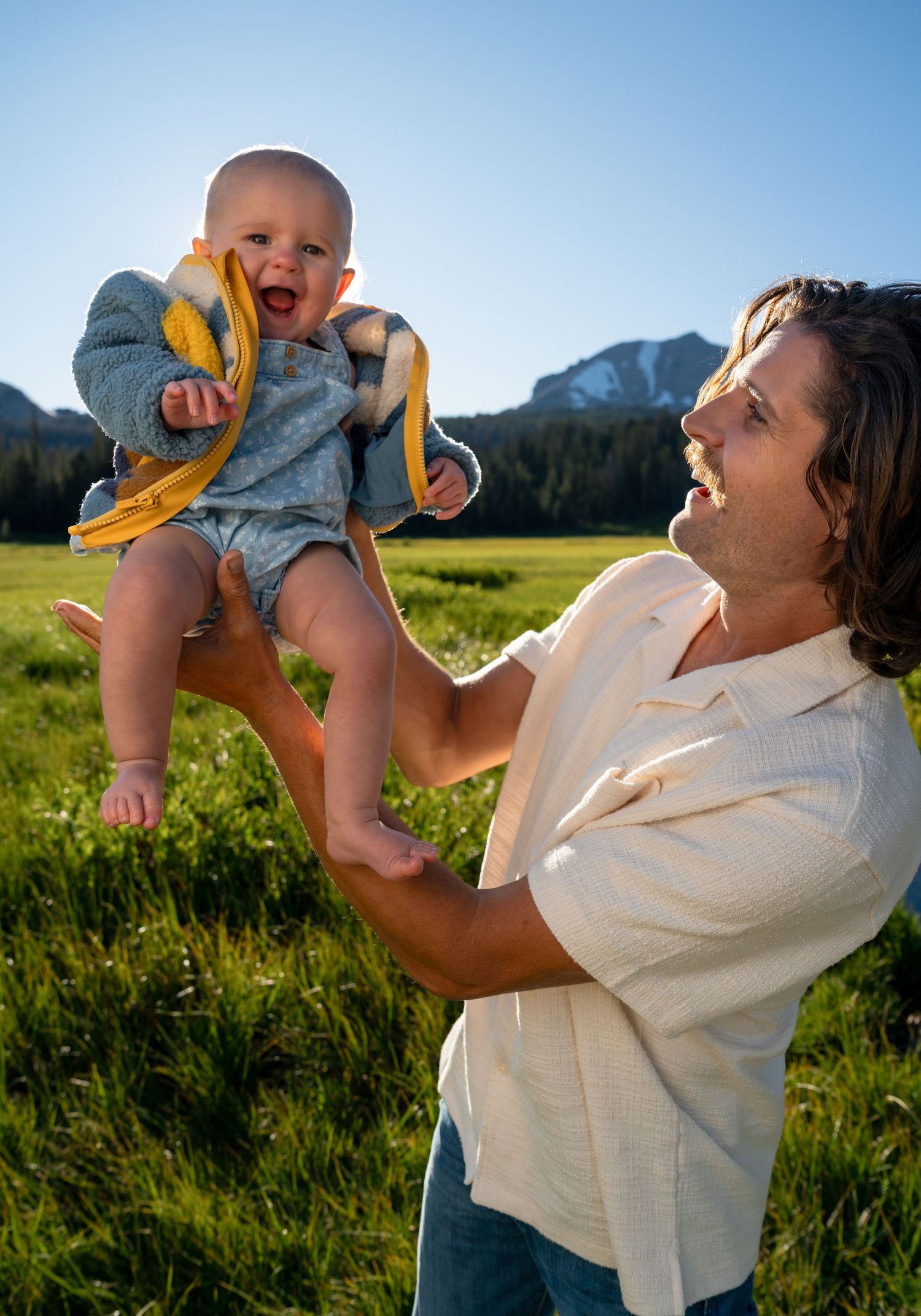 A joyful father lifts his laughing baby outdoors in a sunny green meadow with mountains in the background.
